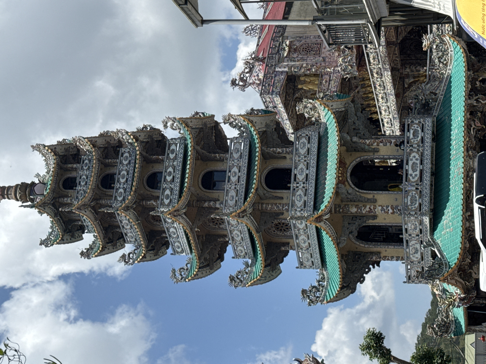 The image shows a tall, ornate pagoda with multiple levels and green tiled roofs. The pagoda is decorated with intricate carvings, including dragons and other mythical creatures. It appears to be a sunny day with a partially cloudy sky. To the right of the pagoda, a portion of a modern building with a metal railing is visible. The pagoda is located in Xuân Thành, Vietnam.