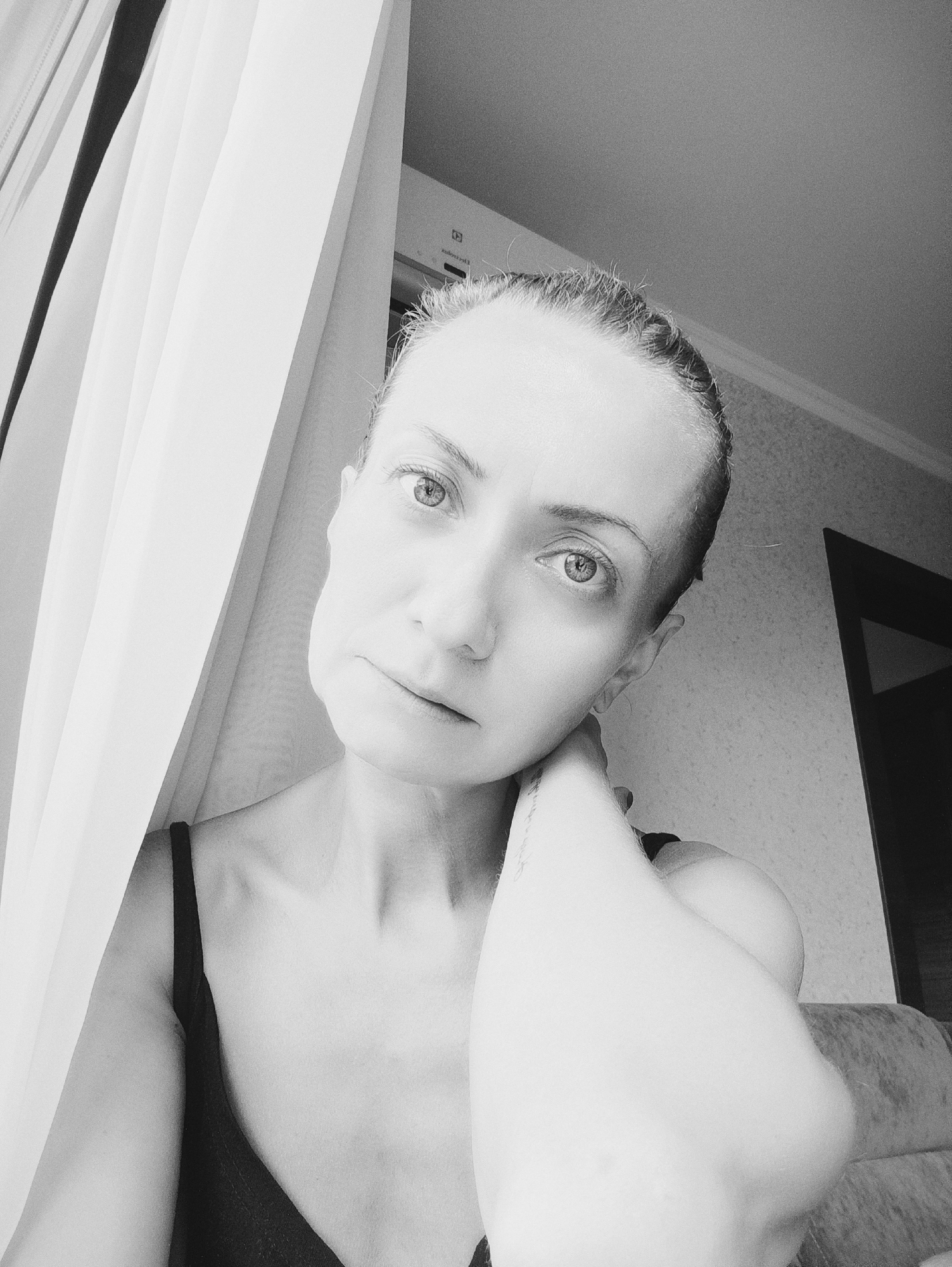 A black and white, medium shot, waist-up selfie of a woman with light hair pulled back and a black camisole. She is positioned to the right of a white curtain, with her left arm raised and her hand touching her neck. Her gaze is directed towards the camera. The background appears to be an indoor setting, possibly a bedroom, with a portion of a doorway visible. There are no discernible landmarks indicating a specific location within Moscow, Russia.
