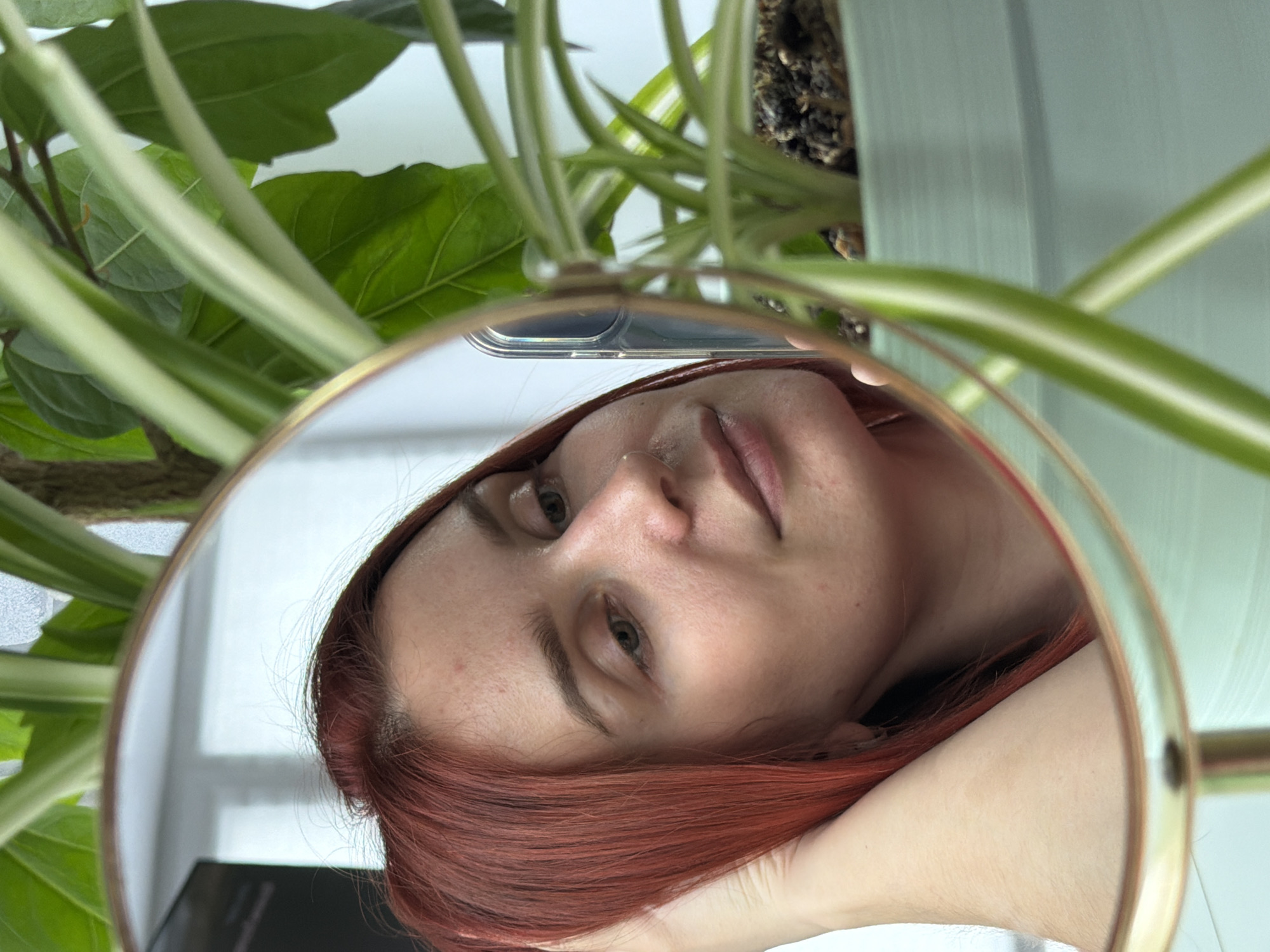 A reflection in a round, gold-rimmed mirror shows a woman with auburn hair, light skin, and green eyes. The top of the mirror reflects a smartphone, green leaves and stems of a plant, and a pale green planter. The woman’s head is resting on her hand, and she is looking at the camera. The background is indistinct, appearing to be a white wall. There is no information to suggest that the image was taken in Chaikovsky, Russia.