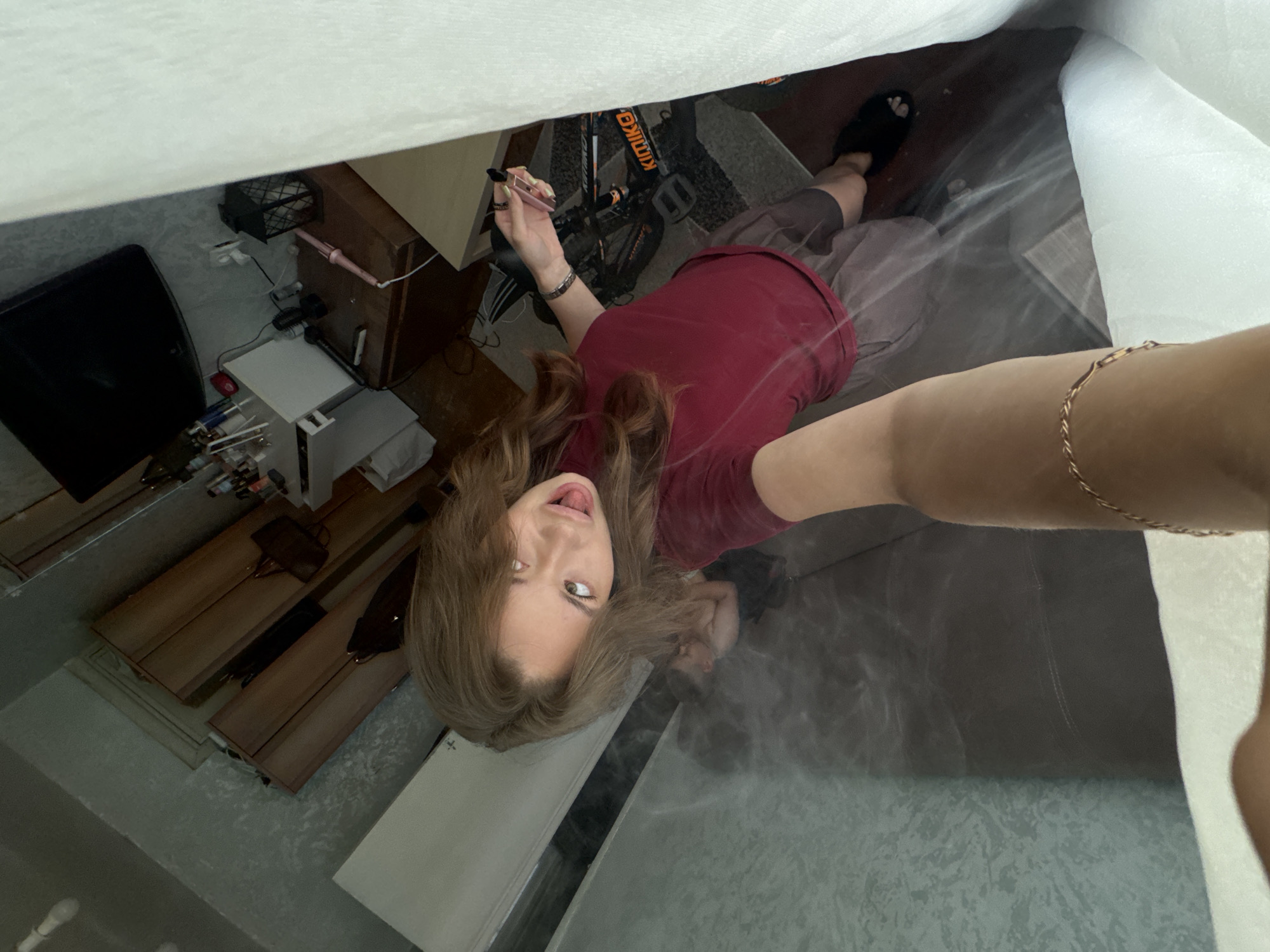 A young woman with long brown hair is taking a selfie from a high angle. She is wearing a maroon t-shirt and gray shorts. Her tongue is sticking out and her eyes are wide open, looking upwards. In the background, a man is visible lying on a couch. The room appears to be a living space with furniture and personal items. The reflection on the floor suggests the presence of water or a reflective surface, possibly a pool or a glass table. The overall lighting is dim, with a white curtain on the right side of the frame.