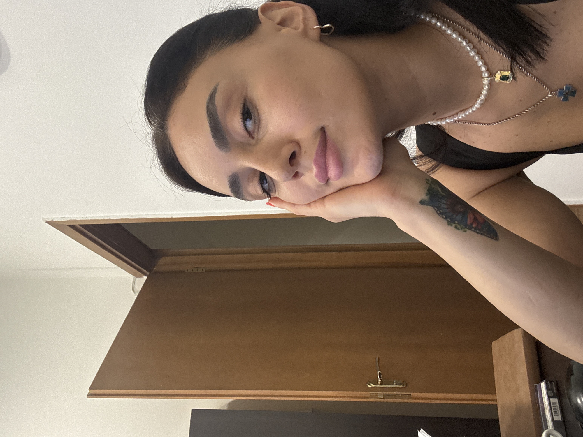 A young woman of Mediterranean or Middle Eastern descent with dark hair and expressive eyes is captured in a close-up selfie. She is resting her right cheek on her left hand, her gaze directed slightly off-camera. Her lips are painted with a soft pink hue, and she wears a delicate pearl necklace with a pendant, layered with another chain necklace featuring a blue cross pendant. A small heart-shaped earring adorns her left ear. On her left forearm, a tattoo of a butterfly is visible. She is wearing a black top.
The background suggests an indoor setting. To the left, a partially open wooden door with a brass handle is visible. The walls are painted a light, neutral color, possibly white or cream. To the far left, a dark rectangular object, possibly a piece of furniture or electronic equipment, is partially visible. In the foreground, on a wooden surface, a stack of what appears to be books or pamphlets and a mouse are present. The lighting is soft, suggesting ambient indoor light. There are no explicit indicators of the specific location within Tbilisi, Georgia, beyond it being an interior space.