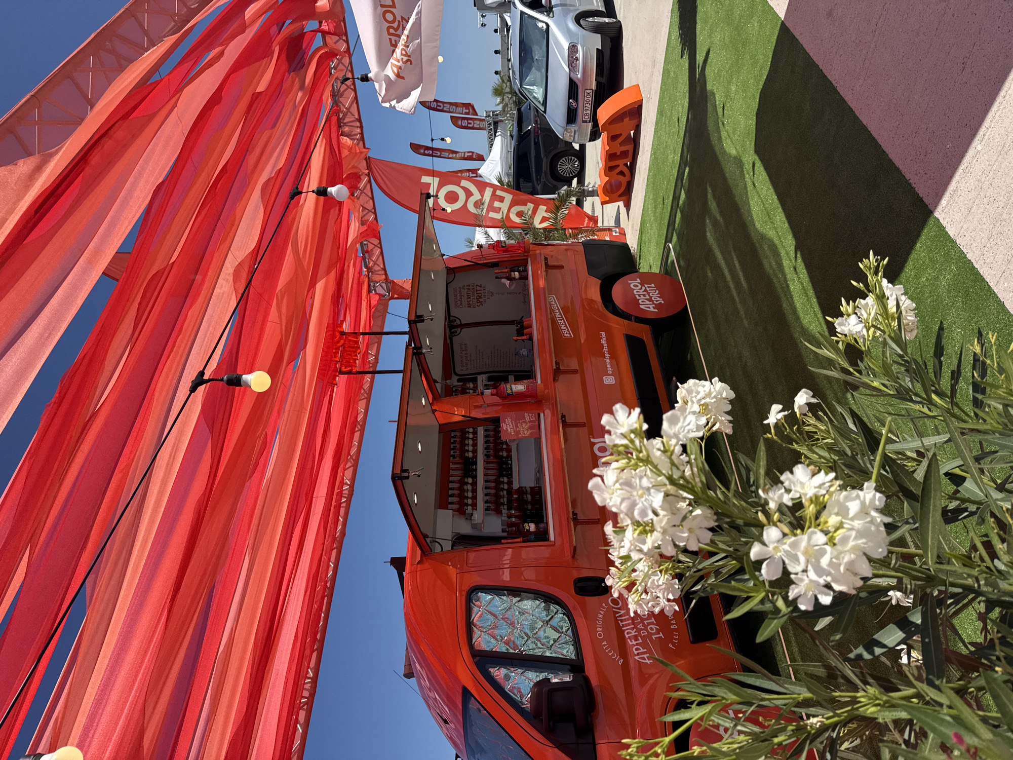 An outdoor Apero Spritz pop-up bar is set up on a sunny day in Pomorie, Bulgaria. The bar is a vibrant orange van with its serving windows open, displaying bottles of Aperol and glasses. Red fabric drapes are strung overhead, creating a festive canopy, and strings of lights are visible. In the foreground, white flowers with green leaves add a touch of natural beauty. Artificial turf covers the ground around the van, and in the background, several cars are parked, including a silver Volkswagen. A large orange sign with the word "APEROL" is also present. The overall scene suggests a lively event or promotional activity.