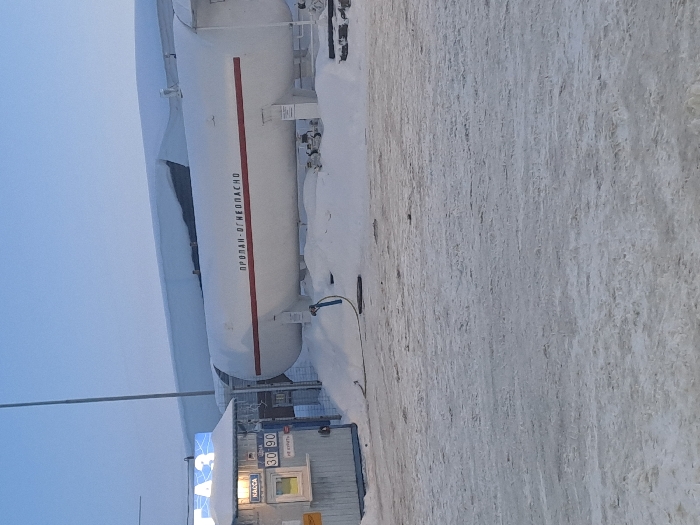 A gas station on a snowy landscape is captured on a dreary day. A large, white propane tank with red lettering dominates the frame. The word “ПРОПАН-ОГНЕОПАСНО” is written on the tank. A small building with a “KACCA” sign is visible beneath the tank. The ground is covered with snow, and the sky is pale and overcast. The overall setting seems desolate and cold.