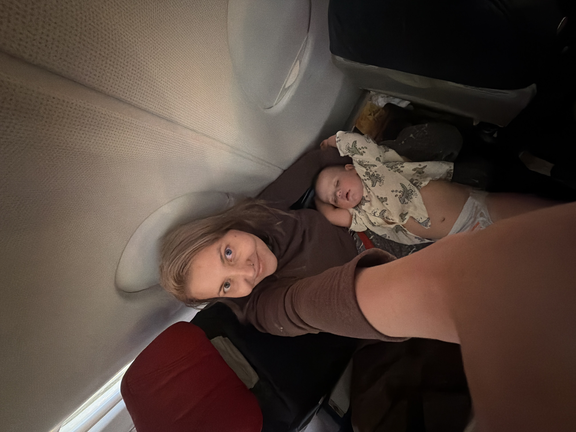 A woman and a baby are in an airplane. The woman is facing the camera, and the baby is sleeping on her lap. The airplane window is visible in the background. There is a red suitcase and other black luggage visible. There is no visible evidence to suggest this image was taken in Da Nang, Vietnam.