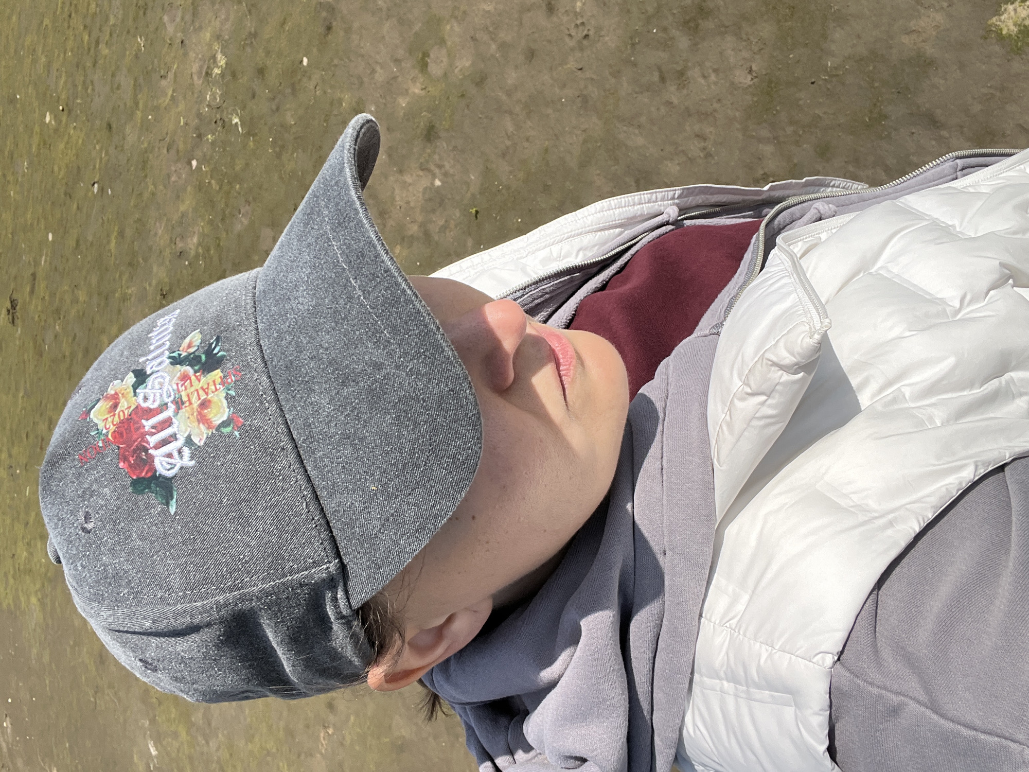 A person, likely female, is shown from the chest up, wearing a gray baseball cap with a floral design and white text that reads "All Saints 2022". They are also wearing a maroon shirt under a gray hoodie, which is then covered by a white puffer vest. The person is looking down and to the right, with their face partially obscured by the cap's brim and shadows. The background is a muddy, unpaved area, possibly a field or shoreline, with some sparse vegetation. The lighting suggests it is daytime. The location is identified as Tullycullion, Ireland.