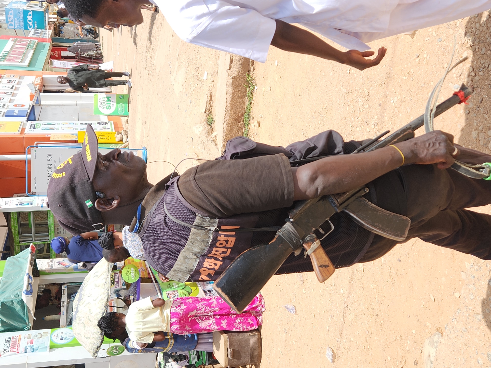 A medium shot captures a man walking in an urban environment. He wears a black baseball cap with a Nigerian flag patch, a dark brown shirt, and a vest with Korean text printed on it. The man is carrying a rifle and a set of rods with what appears to be a rope tied to them over his shoulder. Several shops line the street, displaying signs for telecommunications companies and phone models. People are seen engaging in activities in front of the stores. The ground is a light tan color, indicating an unpaved or sandy surface.