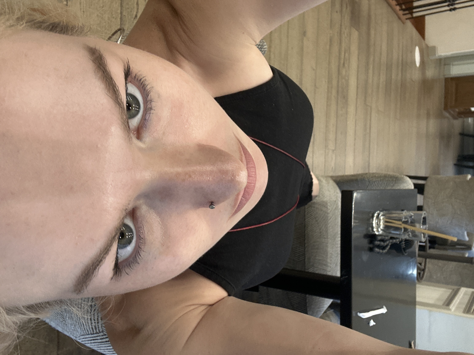 This is a high-angle, close-up, selfie-style photograph of a person's face and upper body. The person is lying down or in a reclined position, looking directly into the camera. The setting appears to be an indoor public space, possibly a cafe or restaurant, with wooden flooring and furniture. In the background, a black table holds a glass with a straw and a small white object, likely a sugar packet or napkin. The person is wearing a black tank top. A nose ring is visible. The lighting suggests an indoor environment, possibly with natural light coming from a window. The image is sharp and clear, capturing the details of the person's facial features and the immediate surroundings. No specific landmarks or indications of Tbilisi, Georgia are present in the image.