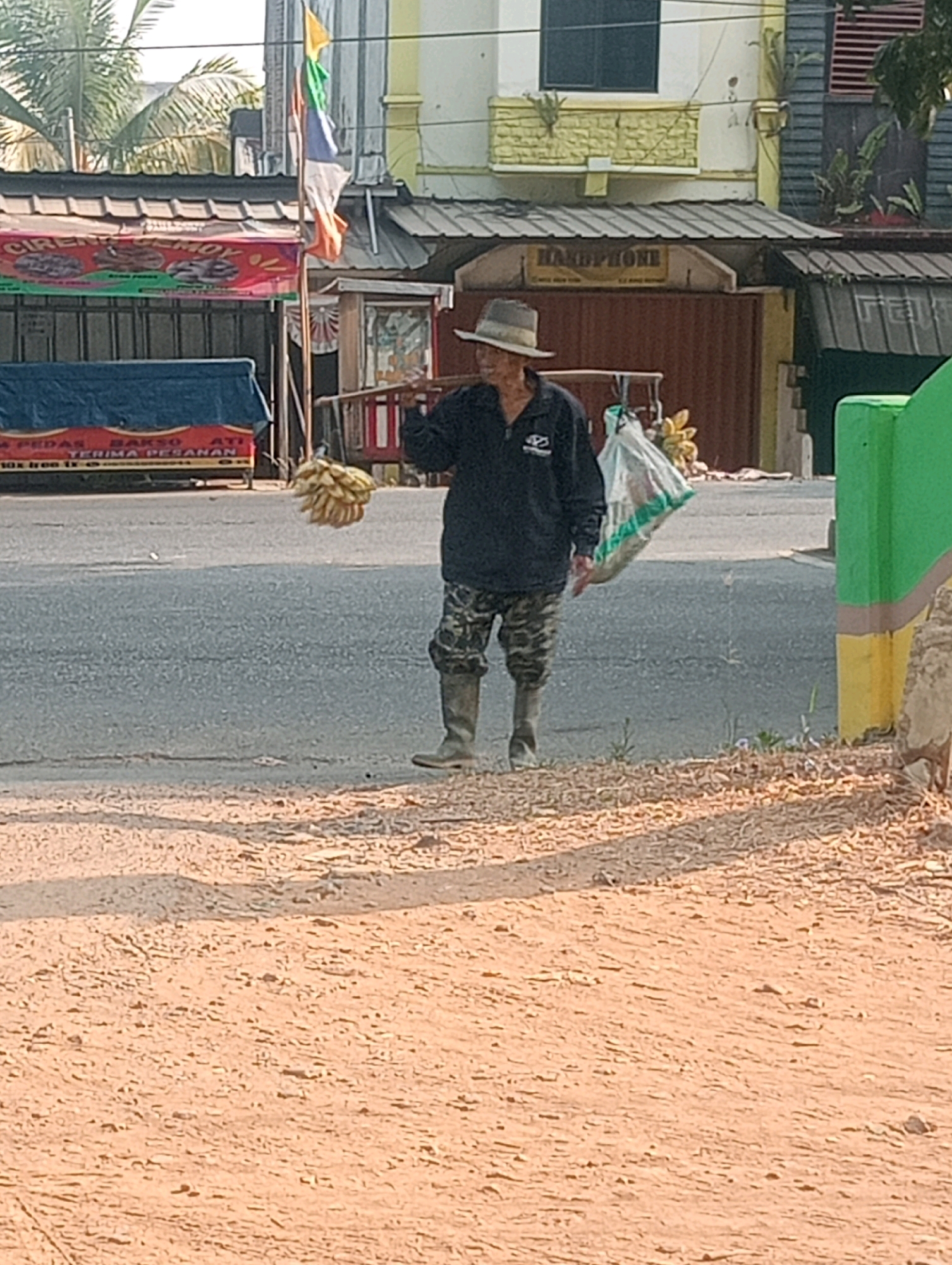 A man wearing a hat and camouflage pants walks across a dusty, sun-drenched road in Bandar Lampung, Indonesia, carrying a yoke over his shoulders with a bunch of bananas on one side and a large white bag on the other. The man is walking from left to right, and appears to be a vendor selling produce. Behind him, a building with a red awning is visible, along with a parked truck. The sky is a clear blue, and the sun casts long shadows on the road. The overall impression is one of a busy, bustling marketplace.