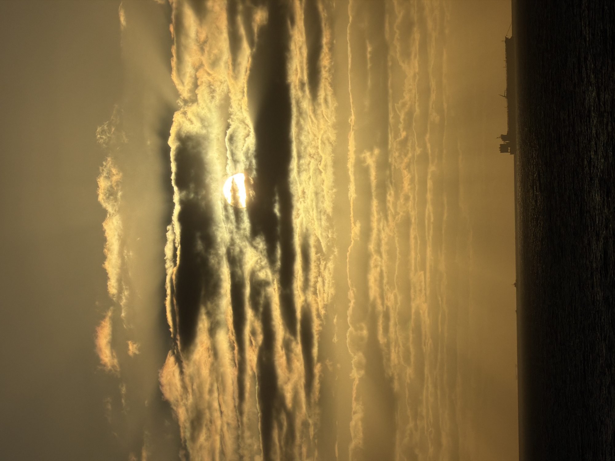 A wide-angle, medium shot depicts a dramatic sky dominated by rows of striated clouds in a muted palette of gold, brown, and grey. The sun, a bright orb, is partially obscured by the clouds, casting rays of light through the gaps. To the right, a dark expanse of water occupies a significant portion of the frame, creating a stark contrast. Along the distant horizon, the faint outline of what appears to be a ship or industrial structure is visible. The image is oriented vertically. There are no people visible in the frame. The location is not discernible from the image provided.