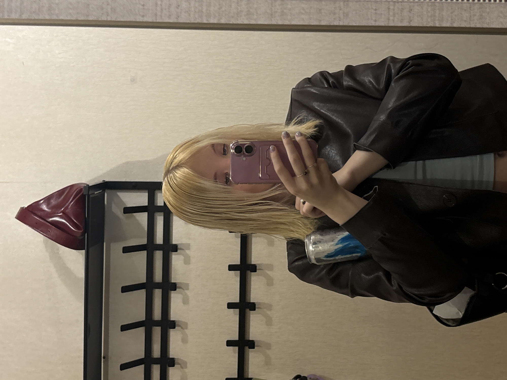 A young woman with blonde hair is taking a mirror selfie in an indoor setting. She is wearing a dark leather jacket over a light blue top. She is holding a pink cellphone in her right hand, with her left hand resting on the phone. Two rings are visible on her fingers. Her hair is long and blonde, falling past her shoulders. In the reflection of the mirror, a rack with horizontal black bars is visible behind her, and on the left side of the rack, a shiny red object, possibly a purse or a decorative item, is placed. To the right of the woman, a silver can with a blue and white pattern is partially visible. The background wall has a textured, light-colored surface. The lighting suggests it could be daytime or artificial light is being used. No text is visible in the image.