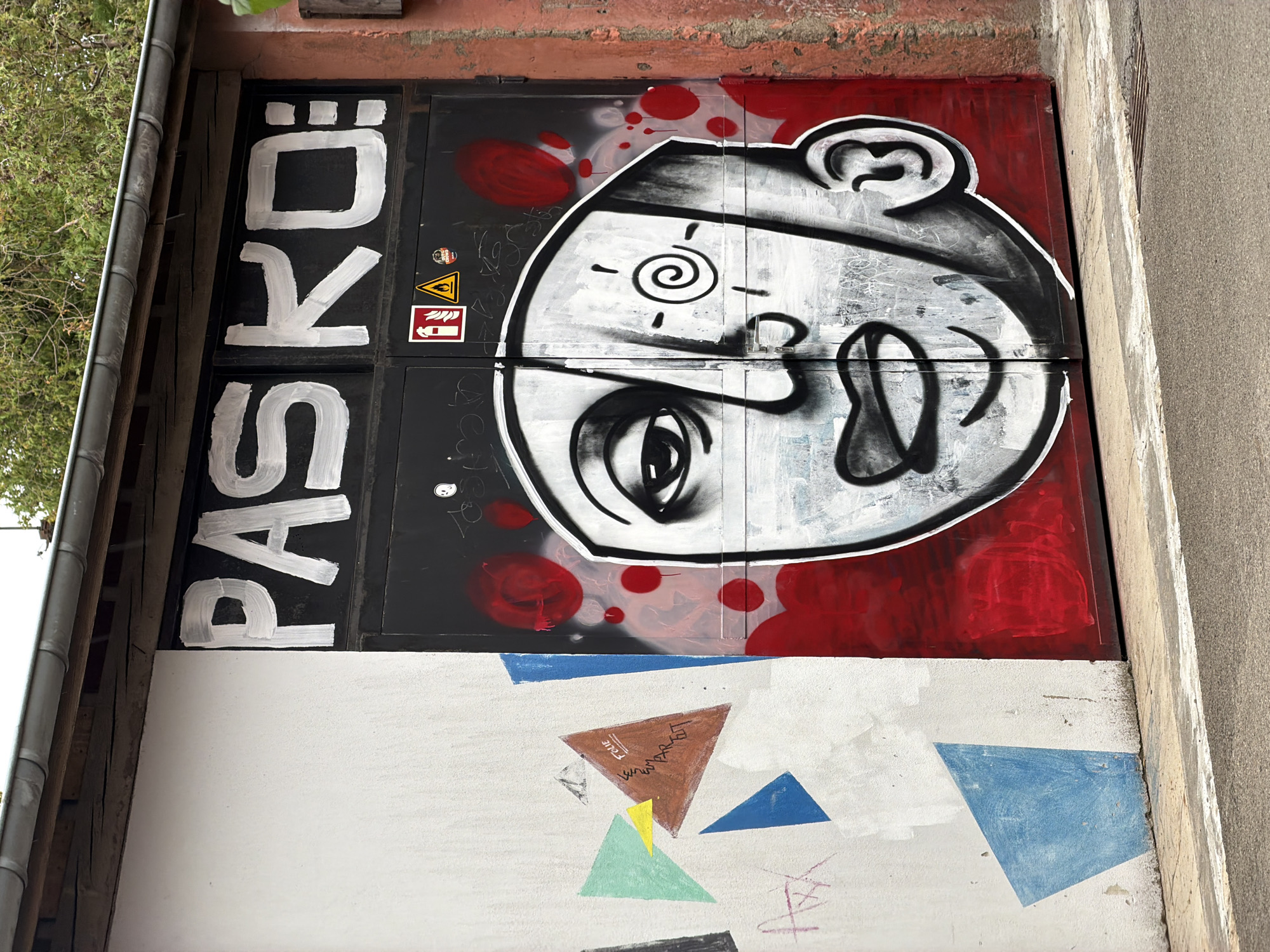 The image displays a close-up of a large industrial garage door adorned with graffiti art in Esch-sur-Alzette, Luxembourg. The garage door, painted black, features prominent white graffiti lettering that reads "PASKQ:". Below this text, a large, stylized human face is painted. The face is split down the middle, with the left side appearing more detailed and shadowed, and the right side rendered in a lighter, more graphic style. A red spiral is depicted on the right side of the face, within a sun-like symbol. The lower portion of the garage door is covered in a vibrant red spray paint, with scattered red circles, and some white scratch marks.
To the left of the garage door, a white wall is visible, also covered in graffiti. This wall displays a collection of abstract geometric shapes in various colors, including blue, green, yellow, and brown. One brown triangle has black text written on it, appearing to read "FOLIE LE SMARRET". Faint pink graffiti lines are also present on this section of the wall.
On the black garage door, just above the face, are two stickers: a red and white fire extinguisher symbol and a yellow triangular warning sign with a flame inside. A small round sticker with a black and white image is also present.
The scene is outdoors, with the top of the image showing a section of a gray metal gutter and some green foliage from trees overhead, suggesting it is daytime. The ground in the foreground is paved with asphalt, with a white painted line marking the edge of the road. A metal grate is visible on the ground in the lower right corner. The right edge of the image shows a textured, reddish-brown wall, possibly made of brick or concrete. The overall impression is one of an urban environment that has been utilized as a canvas for street art.