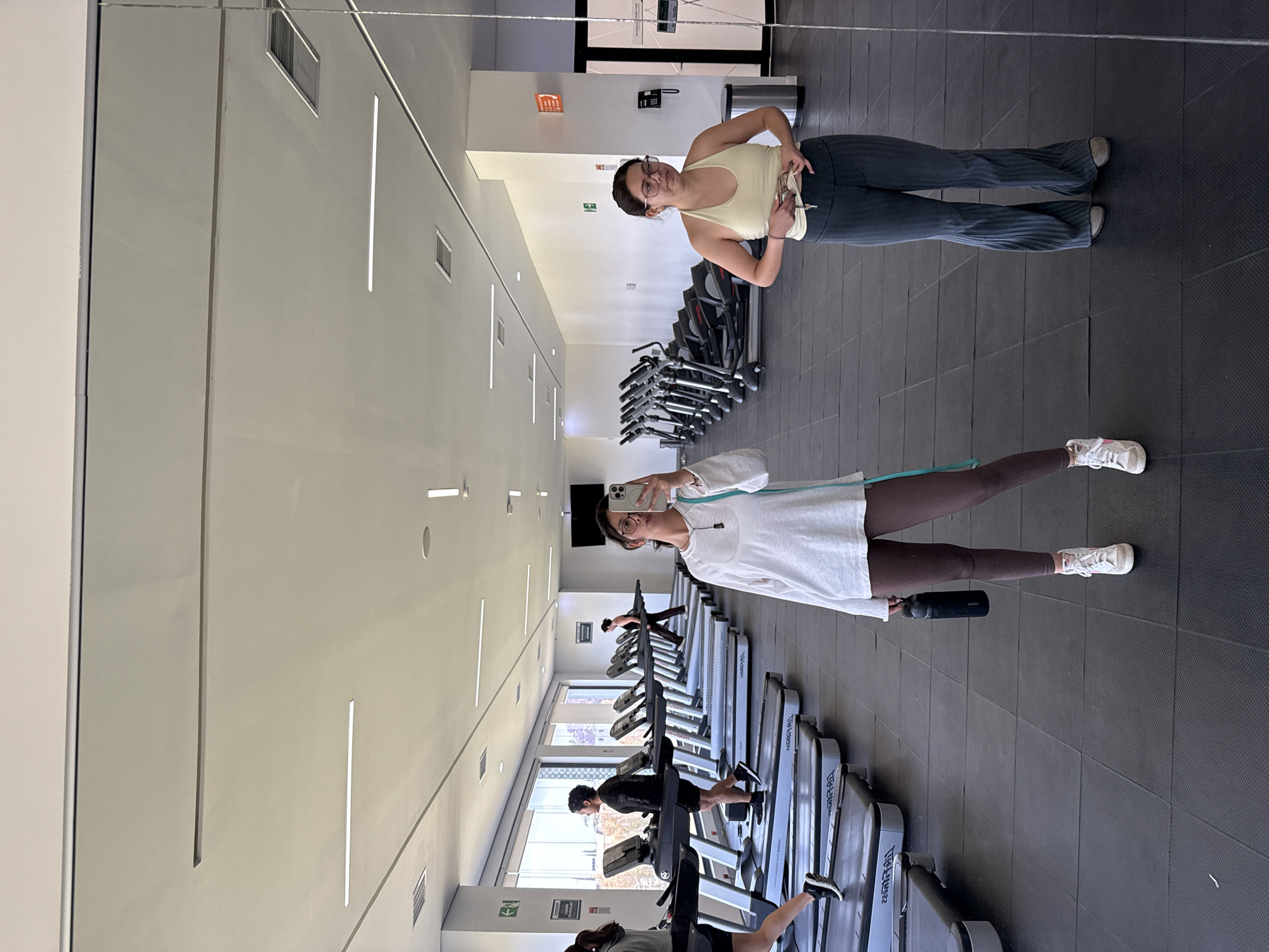 A mirror shot taken in a fitness center in San Andrés Cholula, Mexico, shows a woman in a white shirt, brown leggings, and white sneakers taking a selfie. An exercise band is around her neck, and she is holding a black water bottle. Behind her are rows of treadmills with people exercising. Reflected in the mirror is another woman in a yellow tank top and dark flared pants, standing still with her hands near her waistband. Treadmills and other exercise equipment are visible behind her. The fitness center has a gray tile floor, white walls with linear light fixtures, and exit signs.