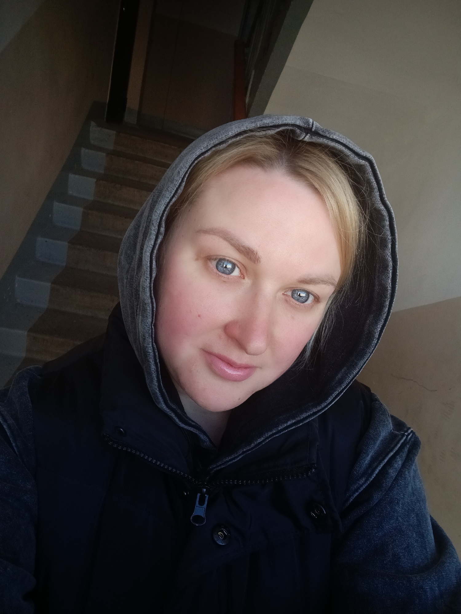 A blonde woman with blue eyes is wearing a hooded jacket. She is standing indoors, with a staircase visible in the background. The location is Chaikovsky, Russia.