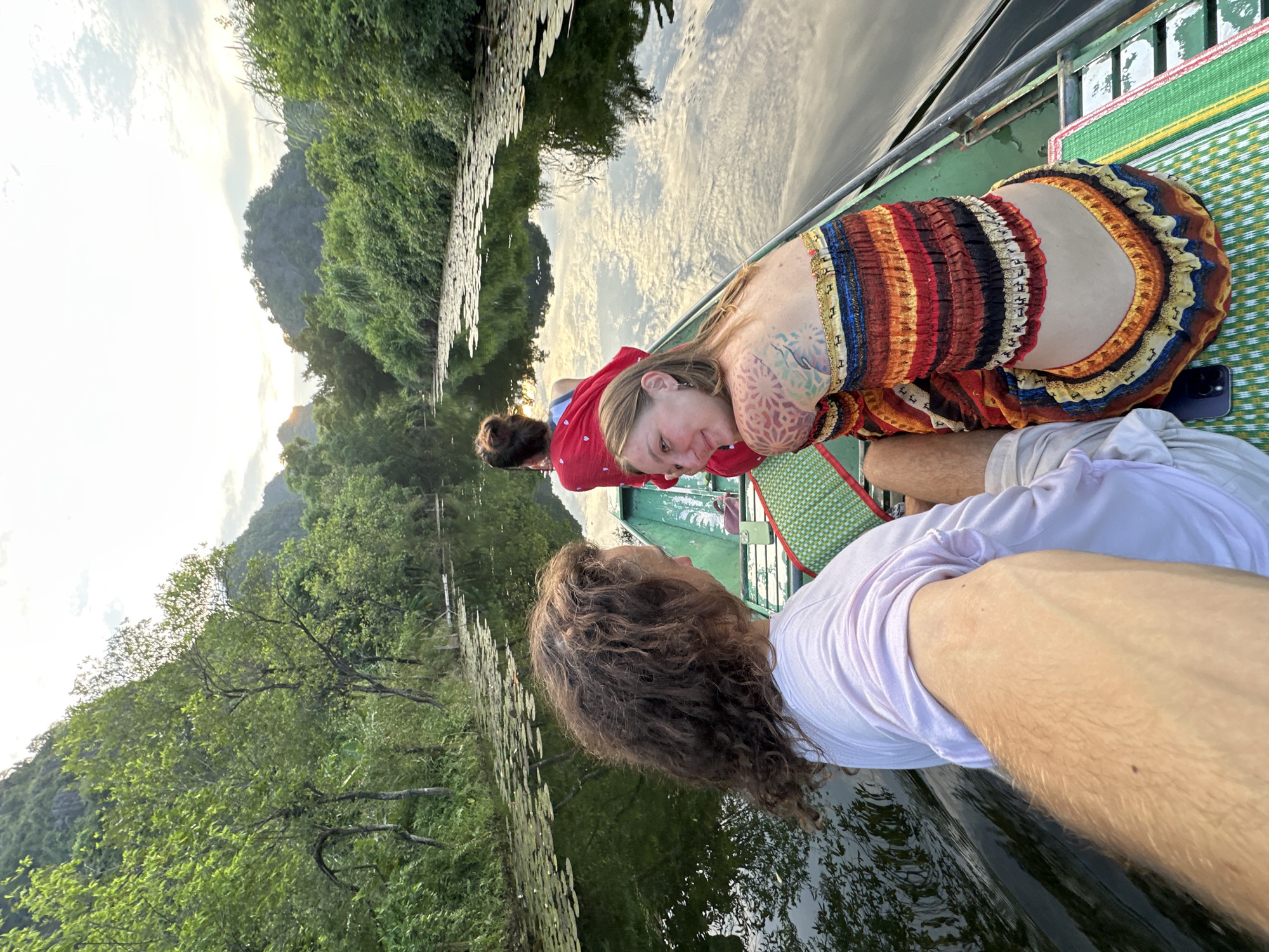 The image shows a group of people on a boat navigating a narrow river surrounded by lush green vegetation and karst mountains. The boat appears to be a traditional Vietnamese longboat, painted green. In the foreground, a man with curly brown hair wearing a pink t-shirt is visible from the chest up, holding the camera. To his right, a woman with blonde hair and a colorful, patterned dress is looking back at the camera and smiling. She has tattoos on her arm and shoulder. Behind them, another person wearing a red top is seated further back in the boat. The river is calm and reflective, with lily pads floating on the surface. The sky is overcast with some sunlight breaking through, casting a warm glow on the scene. The overall impression is one of a peaceful and scenic boat ride through a natural landscape. This scene is characteristic of the Tam Coc area, often referred to as "Ha Long Bay on land," which is located in the vicinity of Hoa Lư, Ninh Bình Province, Vietnam, and part of the broader landscape of Nam Hoa Lư Ward.