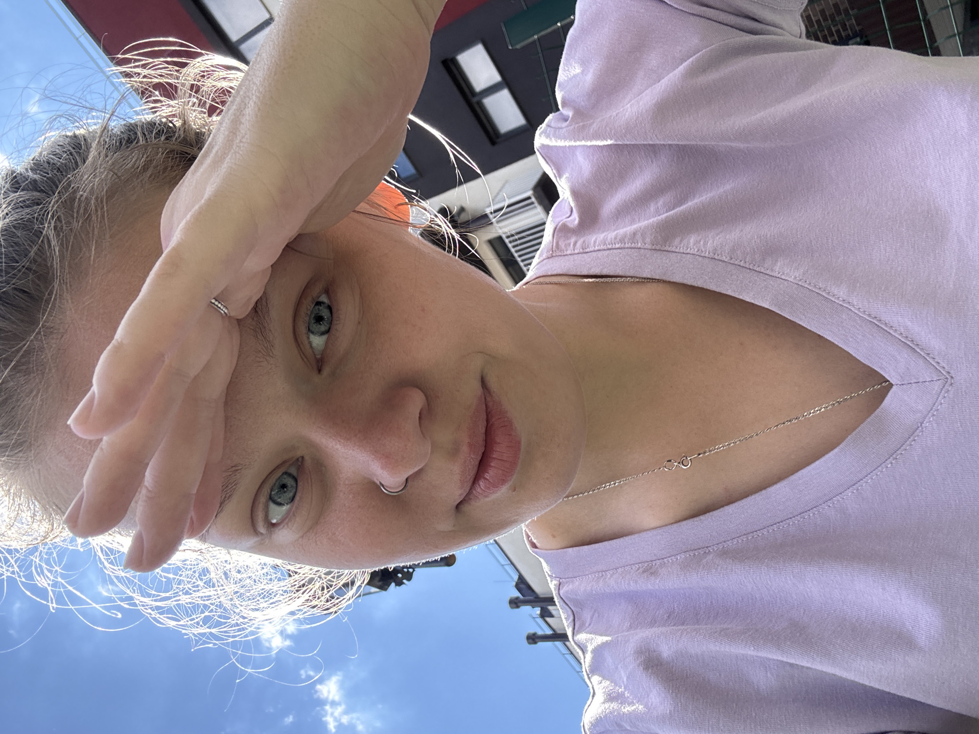 A young Caucasian woman is pictured in an outdoor urban setting, with buildings and a bright blue sky in the background. She is looking directly at the camera with her right hand raised to her forehead. Her blonde hair is pulled back, with wisps of hair illuminated by the sun. She is wearing a light purple V-neck t-shirt and a silver necklace. A nose ring is visible on her left nostril, and a thin silver ring is on her right ring finger. The overall scene suggests a casual, sunny day. The specific location in the city of Vyazniki, Russia, cannot be determined from the visual information provided.