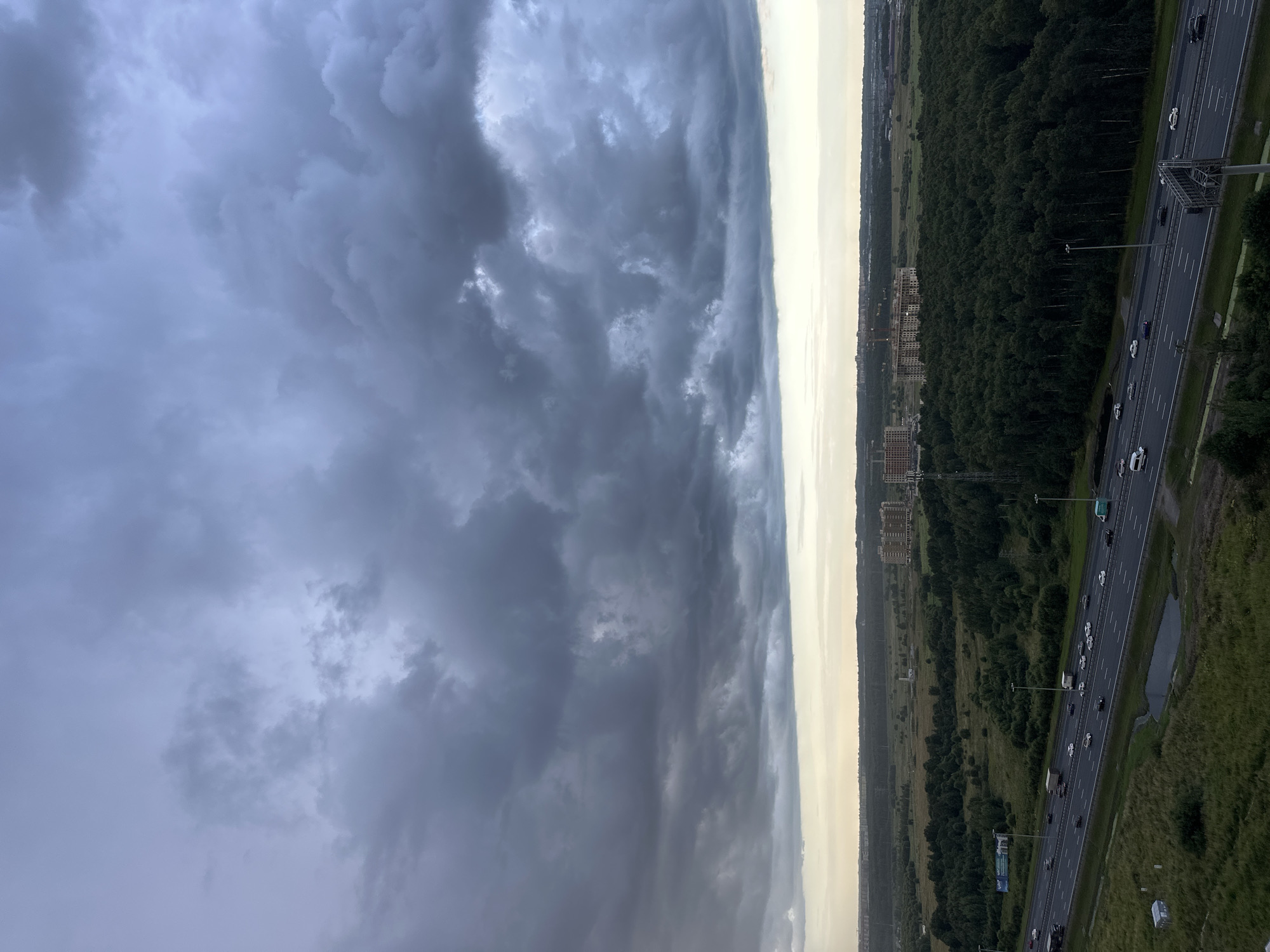 The image depicts a view of a highway with traffic and surrounding landscape under a dramatic, overcast sky. In the foreground, a multi-lane highway stretches across the frame, with numerous cars and trucks visible, moving in both directions. To the left of the highway, there is a grassy area with some low structures. To the right of the highway, a dense, dark green forest dominates the midground. Beyond the forest, a flat landscape extends towards the horizon, featuring scattered buildings, including several tall apartment complexes, some of which appear to be under construction. The sky is filled with heavy, dark gray clouds, suggesting an impending storm or the aftermath of one, with a pale band of lighter sky visible near the horizon. The scene appears to be on the outskirts of a populated area, possibly residential or industrial, adjacent to natural and man-made infrastructure. The location is identified as being in the city of Shlisselburg, Russia.