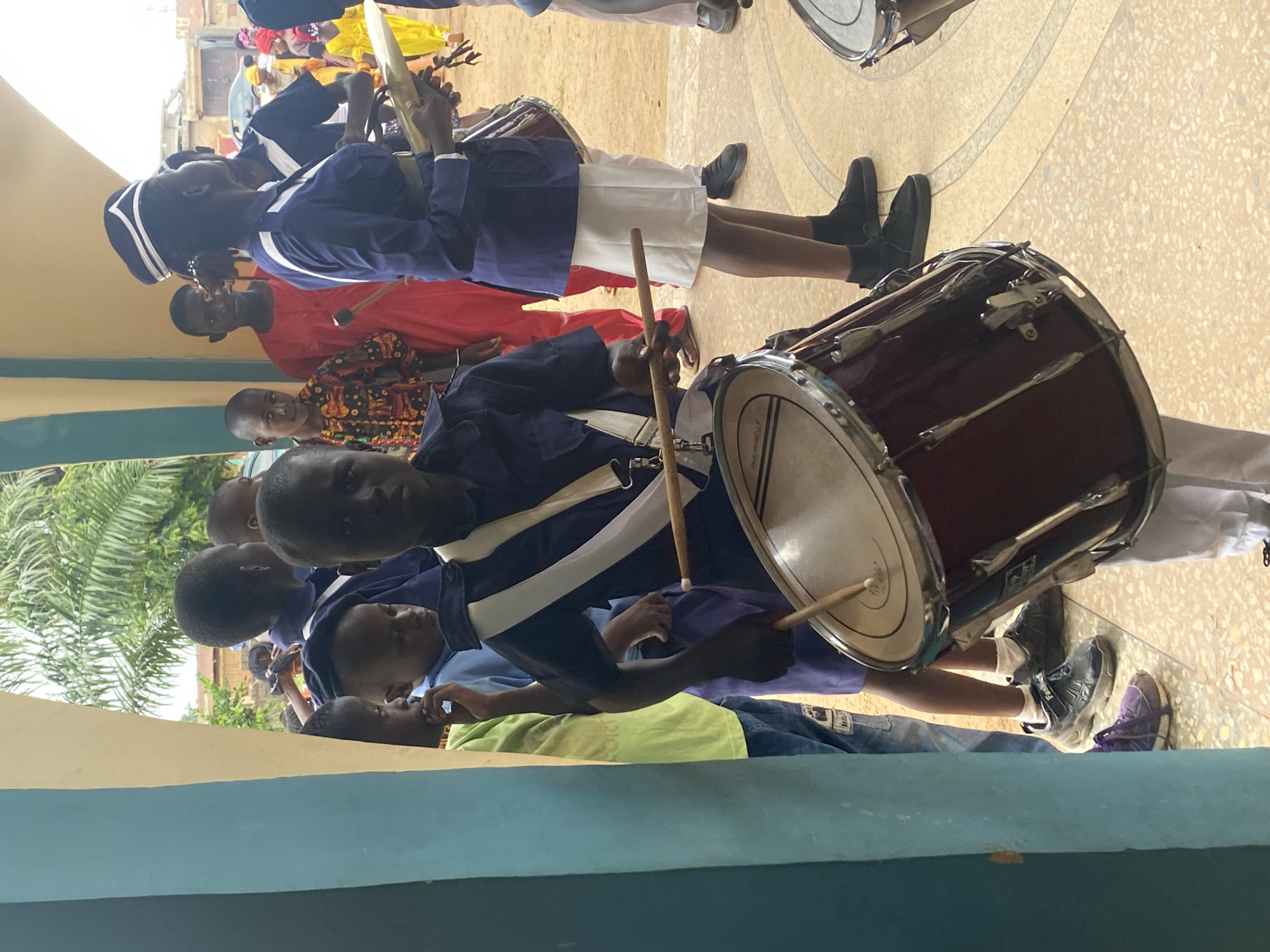 In Kafanchan B, Nigeria, a group of people are gathered in what appears to be an outdoor event. Several individuals are dressed in marching band attire, including navy blue jackets, white accents, and hats. One person is actively playing a large drum with drumsticks. Other people are observing or participating in the event, some wearing colorful traditional clothing. The environment includes building structures, painted walls, and some vegetation.