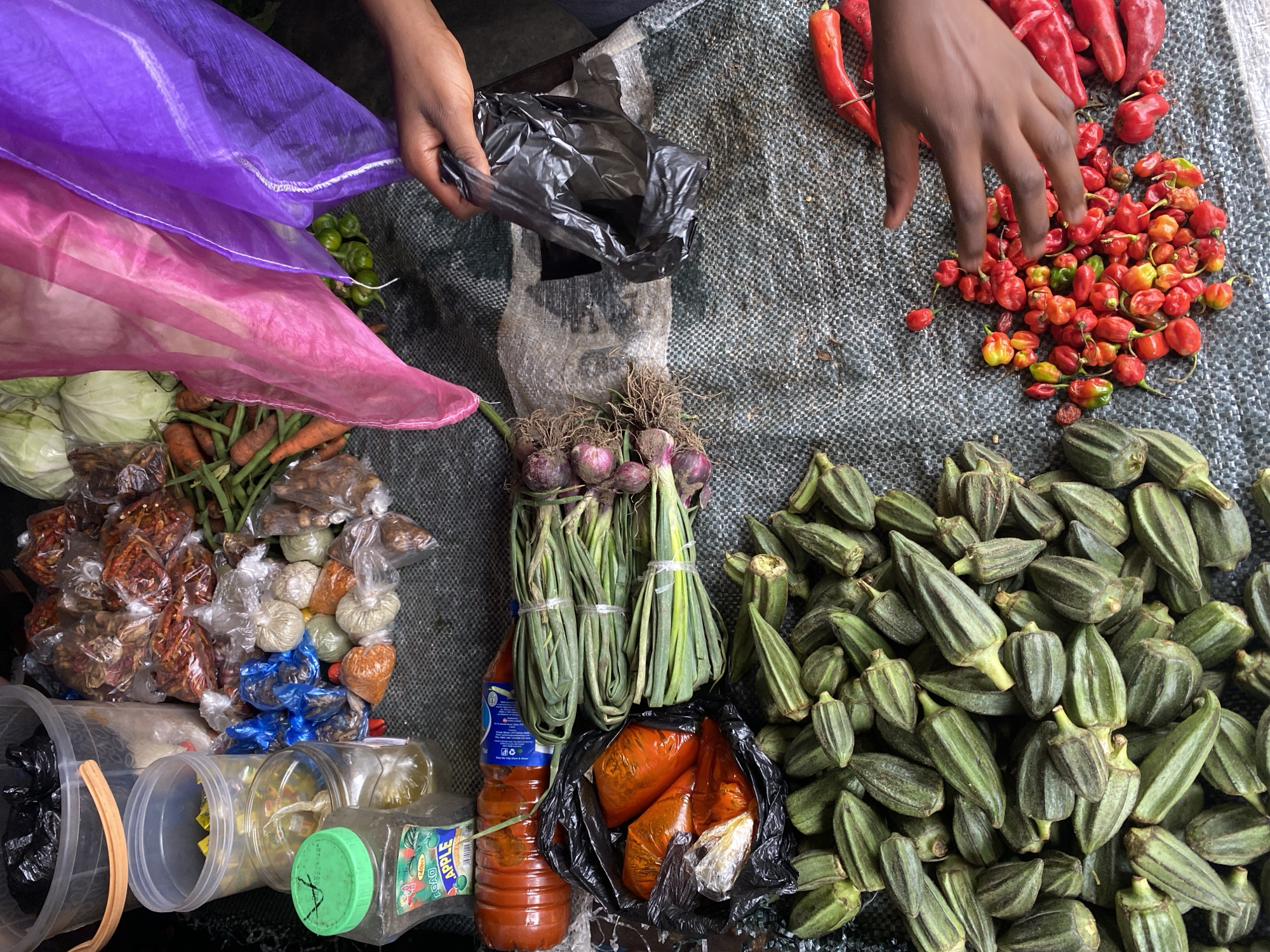 The image depicts a market stall in Gura Top, Nigeria, displaying a variety of fresh produce and packaged goods. In the foreground, a large pile of green okra is visible, with a hand reaching for a cluster of small, colorful chili peppers. To the right of the okra, a bunch of red chili peppers is also present. Behind the okra, a collection of red onions with their roots attached is laid out. Further back, a bunch of green onions or scallions is bundled together, lying next to a bottle of what appears to be chili sauce.
In the upper portion of the image, various packaged spices and dried goods are arranged, some in clear plastic bags and others in larger containers. Among these are small packets of reddish-orange spices and what look like dried chilies. A large transparent bucket with an orange handle is at the top left, containing more packaged items. Two large pieces of sheer fabric, one pink and one purple, are draped over some of the produce, possibly to shade them. The overall setting is that of an open-air market with a rough, textured surface serving as the market stall's display area. The lighting suggests it is daytime.