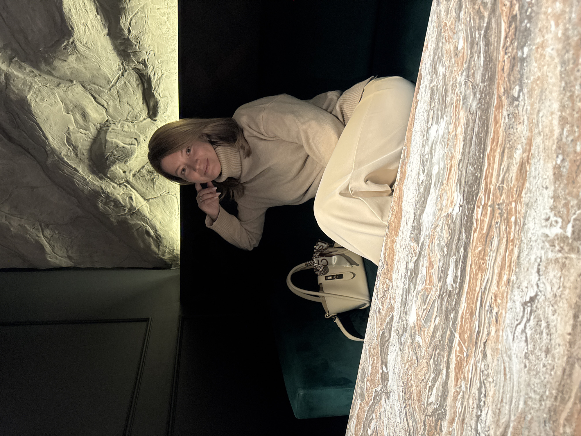 A woman is lying on a dark green velvet couch, wearing a light beige turtleneck sweater and light beige pants. She has her head propped up with her hand, looking at the camera. Next to her is a light beige purse with a decorative scarf tied to the handle. The setting appears to be indoors, with a faux rock wall on one side and a faux marble wall on the other. A dark door is visible behind the faux rock wall. The location is Chaikovsky, Russia.