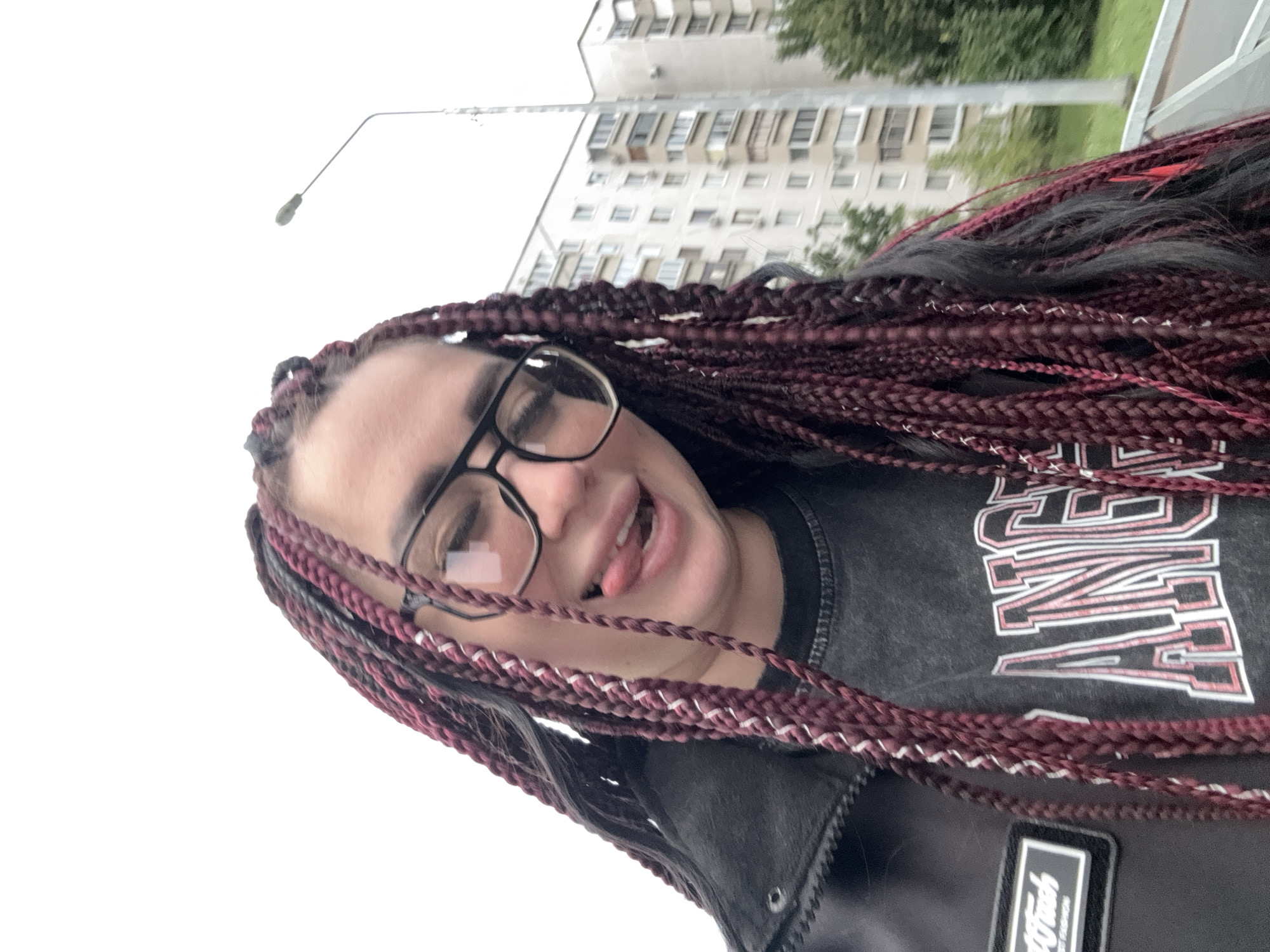 A young woman with burgundy and black braided hair, wearing glasses and a black jacket over a dark t-shirt with a white and pink graphic, sticks her tongue out at the camera. She is outdoors, with a tall, light-colored apartment building and trees in the background. A street lamp is visible on the right side of the image. The scene appears to be a residential area in the city of Tolyatti, Russia.