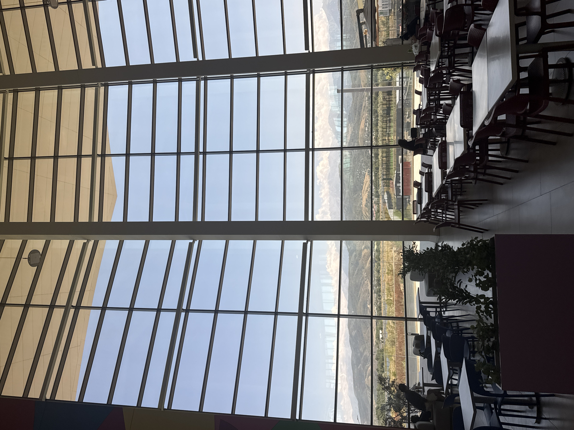 The image shows the interior of a modern, spacious building with a large, curved glass wall that offers a panoramic view of a landscape with mountains in the distance. The interior features a dining area with numerous tables and chairs. The tables are rectangular and some are circular, with light-colored tops and dark bases. The chairs are dark, likely made of plastic or metal, and are arranged around the tables. In the foreground, there is a large planter box with green plants. Several people are visible in the dining area, some sitting at tables and others walking. The overall lighting suggests it is daytime, with sunlight streaming through the glass wall. The architectural style is contemporary, characterized by clean lines and extensive use of glass. The view outside the glass wall depicts a dry, arid landscape with rolling hills and snow-capped mountains in the background. There is also a body of water visible in the middle distance. This scene is likely located in the city of Almaty, Kazakhstan, given the characteristic mountainous backdrop.