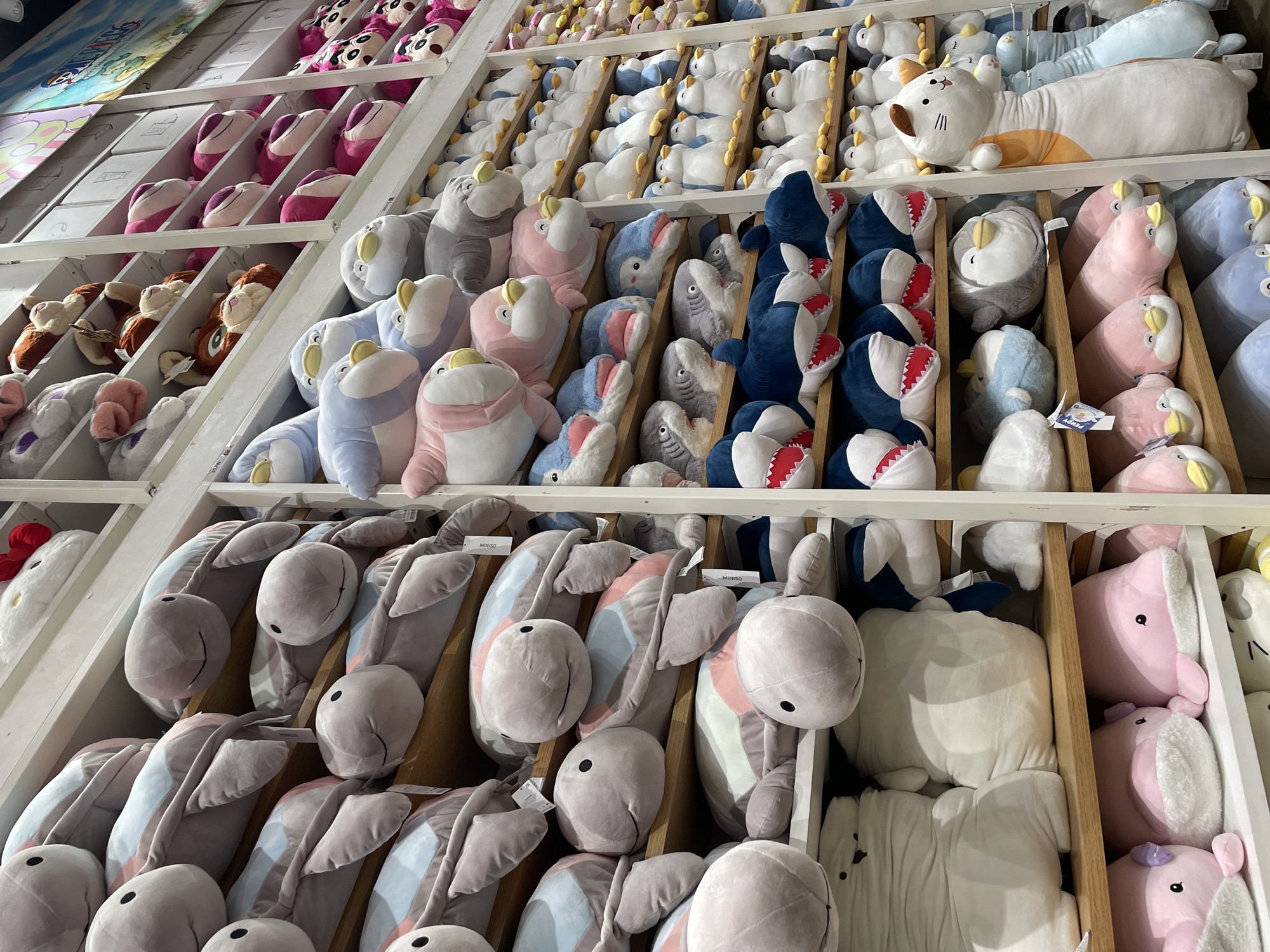 This is a close-up, eye-level shot of shelves filled with an assortment of plush toys in what appears to be a retail store. The shelves are made of wood and the overall color palette is soft, with dominant colors of light grey, pink, blue, and white.
On the left side, the shelves are stacked with large, grey plush turtles with pastel blue and pink shells, each with simple black eyes and a smiling mouth. Below these, there are rows of large white plush toys, some with dark accents. Further down are pink, whale-shaped plush toys with white bellies and small purple eyes.
The center shelves feature a variety of plush animals. There are blue and grey penguins with yellow beaks, some with pink accents. These are interspersed with plush sharks with open mouths revealing white teeth. Other toys visible include what appear to be seals or other marine mammals, and some white, fluffy objects.
The right side of the image shows more shelves stocked with plush toys, including rows of white penguins, and a very tall, white and orange cat-shaped plush toy standing upright. In the background, there are glimpses of other merchandise and signage, including a brightly colored mural or poster featuring cartoon characters.
The lighting is bright and even, suggesting an indoor retail environment. There are no people visible in the frame. The image is focused on the detailed arrangement and variety of the stuffed animals. The presence of a "MINISO" tag on some of the toys suggests this location is a Miniso store. Based on available information about Miniso stores in the Philippines, a likely location for this particular display could be **SM City Davao, Philippines**.