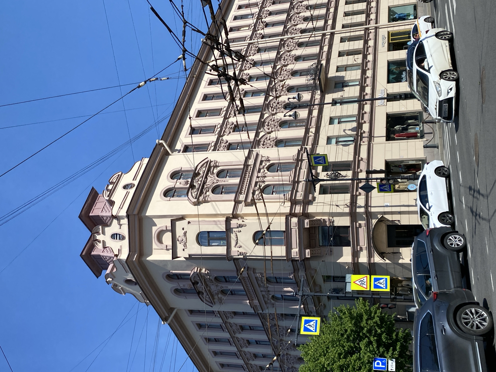 The image presents a bright, sunny street scene in Saint Petersburg, Russia, centered on a grand, multi-story building. The building features an ornate facade with intricate decorative elements around numerous windows and balconies, painted in contrasting cream and pale purple-grey tones. The ground floor houses shopfronts, notably "Versace" and "Max Mara."
Along the street to the right of the building, several vehicles are visible, including a white hatchback with a Russian license plate ("757РМ 98," indicating Saint Petersburg registration) and parts of two dark grey SUVs. A network of black utility wires crisscrosses the clear blue sky, extending across the upper portion of the image.
The scene is illuminated by strong sunlight, suggesting midday or early afternoon. A small, vibrant green tree is visible at the bottom right. Several street signs are present, including multiple yellow and blue pedestrian crossing signs, and a blue "P" parking sign indicating "↖ 25м." No people are clearly visible in the frame. The overall atmosphere is that of a bustling urban environment on a clear, bright day.