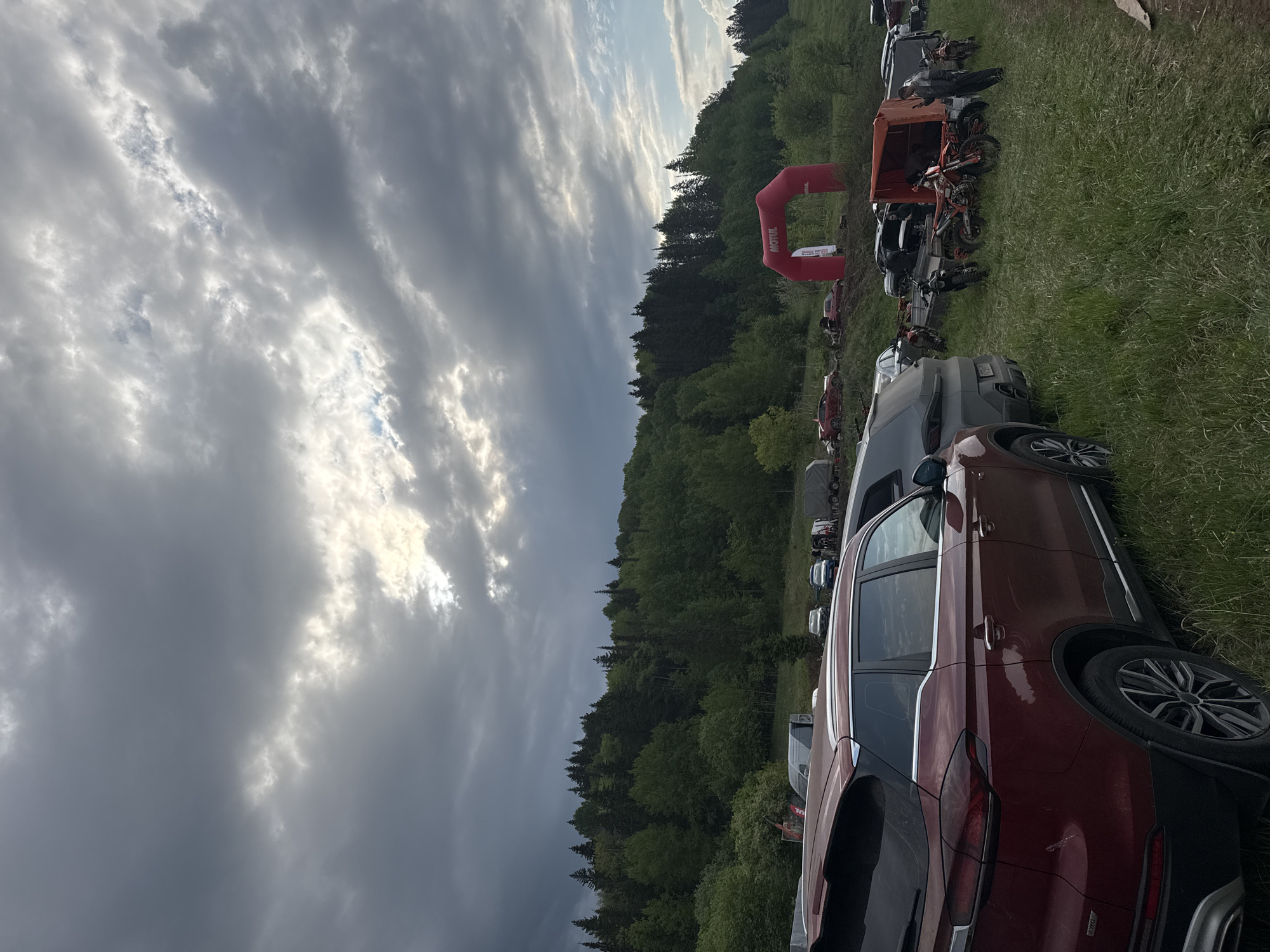 The image depicts a rural scene near Чёрная, Russia, under a cloudy sky with patches of sunlight breaking through. A forest lines the horizon. In the foreground, several vehicles are parked in a grassy field, including a dark red SUV. Further back, there are motorcycles and a red Motul inflatable arch. No people are clearly visible performing actions.