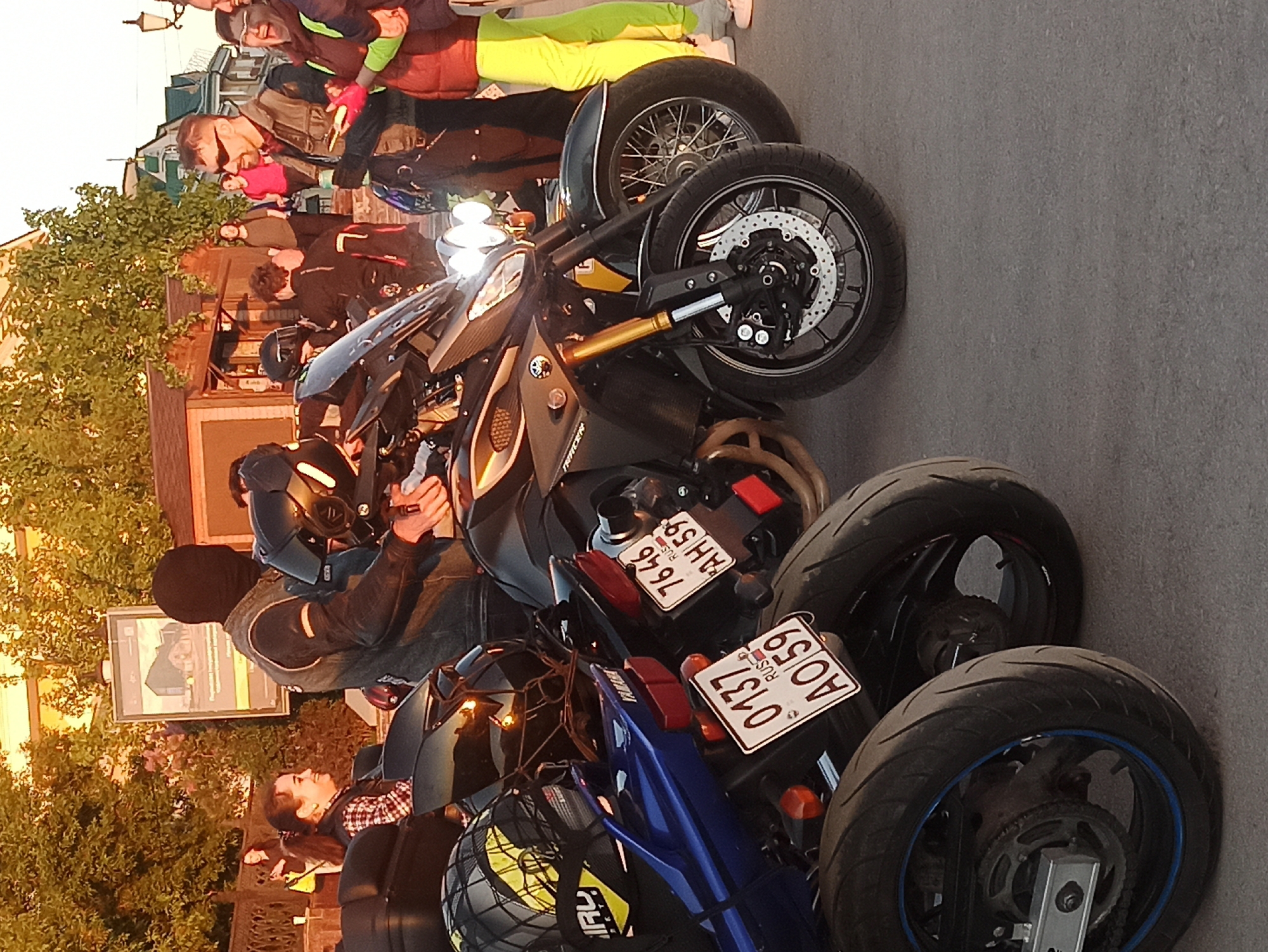 The image shows a street scene in Unknown, Unknown, featuring a group of people standing near a row of parked motorcycles. Three motorcycles are prominently displayed in the foreground. The middle motorcycle is a dark-colored sportbike with its headlight on. The license plates on the motorcycles read "0137 AO 59 RUS" and "7646 AH 59 RUS". A man wearing a black beanie and a leather jacket is seated on one of the motorcycles and appears to be adjusting his helmet. Other people are visible standing and talking in the background. The setting appears to be near a building or structure and some trees are visible. The lighting suggests that it is either sunrise or sunset.