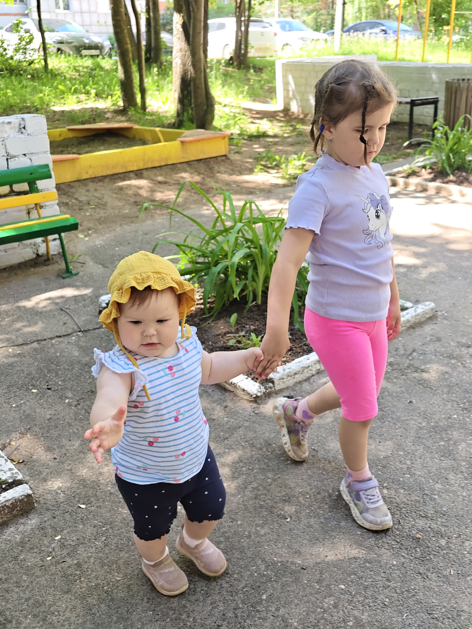 The image depicts two young girls holding hands and walking on a paved surface. The girl on the right, appearing older, has braided hair and wears a light purple t-shirt with a unicorn graphic and pink capri pants. The girl on the left, an infant, wears a yellow sun hat, a striped t-shirt, and dark blue shorts. They are in an outdoor environment, possibly a playground or park, with trees, green foliage, a sandbox, and a bench visible in the background. The overall scene suggests a casual, daytime activity.