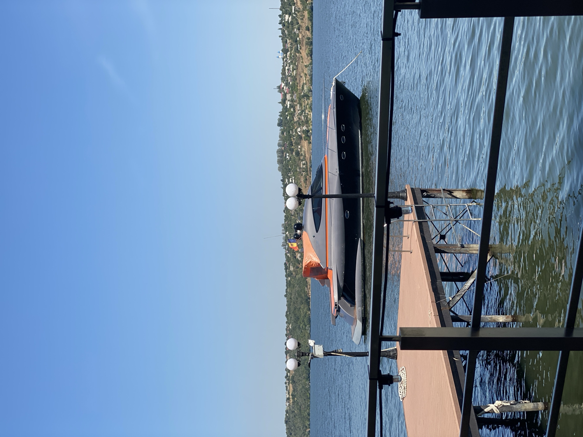 The image captures a serene daytime scene on a body of water, likely a river or lake, with a picturesque backdrop of a verdant, hilly shoreline dotted with buildings. In the foreground, a section of a dock with a brown wooden deck is visible, supported by metal stilts submerged in the water. A black metal railing runs across the bottom and mid-section of the frame, partially obscuring the view of the dock and the water. A tall lamp post with two spherical lights stands on the dock.
Dominating the mid-ground is a sleek, modern yacht with a dark grey hull and striking orange accents on its superstructure. The yacht is moored and floats calmly on the water. A Romanian flag is visible flying from a small mast on the stern of the yacht. The water is calm with gentle ripples, reflecting the clear blue sky and the surrounding landscape. The shoreline features a dense line of trees and vegetation, with houses and structures spread across the hills. The overall impression is one of tranquility and a pleasant, sunny day. While the prompt suggests the location is Ustia, Moldova, this cannot be confirmed solely from the visual information provided in the image.
