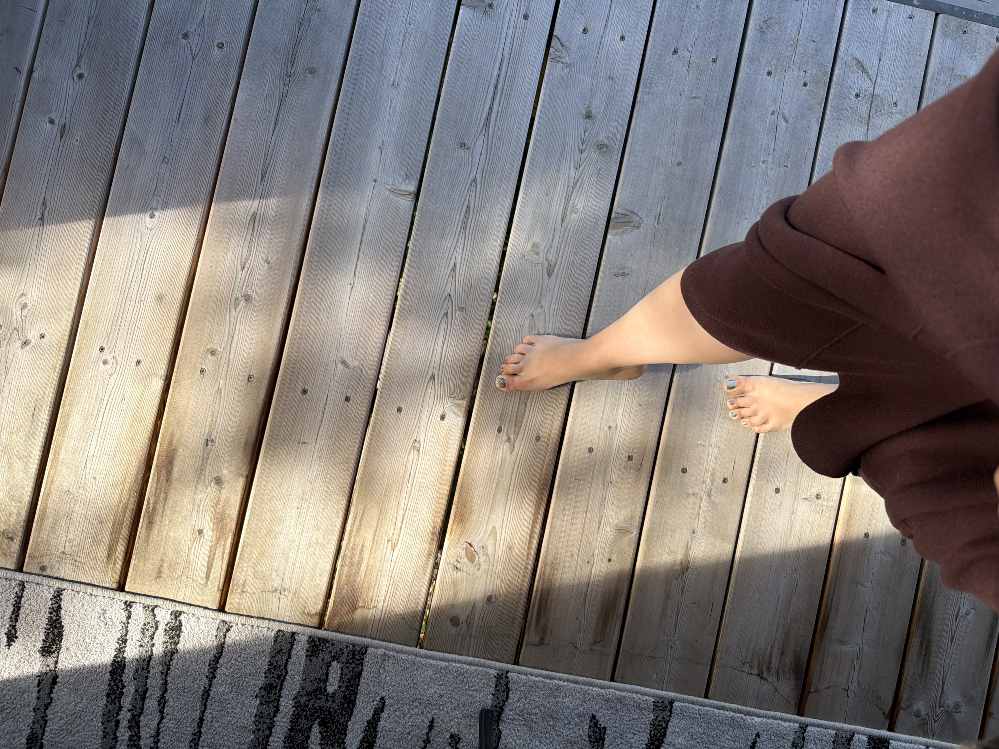 An overhead shot shows a person's feet and legs standing on a wooden deck. The deck is composed of light-colored planks laid horizontally. A shadow cuts diagonally across the deck from the top left to the bottom right, suggesting sunlight. To the left of the deck, a portion of a grey and black patterned rug is visible. The person is wearing a dark brown garment that covers their legs from the knees down. Their feet are bare, and their toenails are painted with a light blue and grey polish. The environment appears to be outdoors, possibly a patio or balcony. The image's focal point is the person's feet against the textured wooden surface.