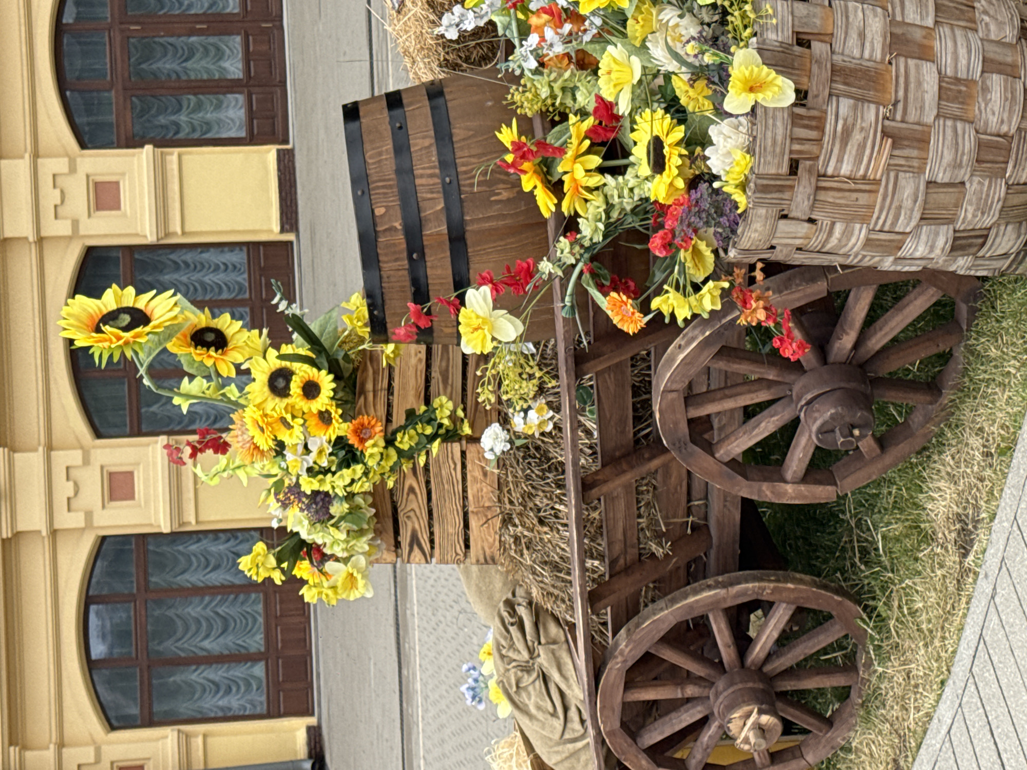 The image depicts a decorative display of a wooden cart filled with hay, a wooden barrel, and an assortment of artificial flowers, primarily sunflowers, yellow daisies, and red flowers. The cart is positioned outdoors on a grassy area with a paved walkway in the foreground. In the background, a yellow building with large arched windows is visible. The scene appears to be a static display, possibly for a seasonal event or decoration. There are no people or animals in the image. The overall impression is one of rustic charm and autumnal or harvest-themed decor.