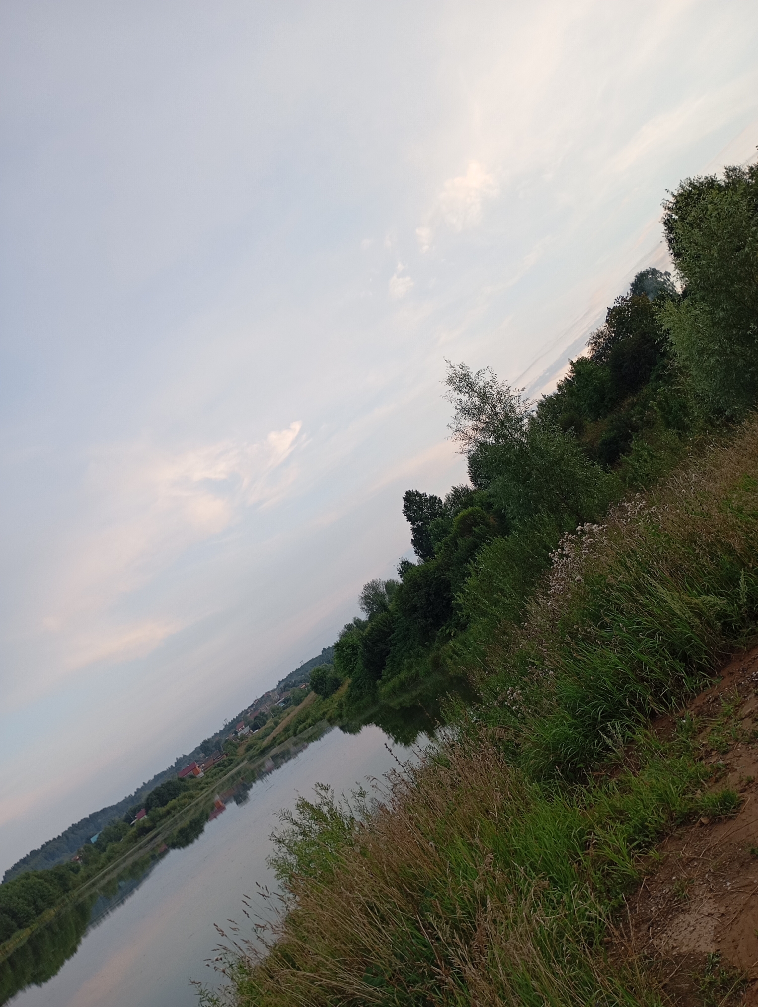 The image depicts a body of water, possibly a river or lake, with a bank covered in dense green vegetation and dry grasses in the foreground. The water reflects the sky and the lush green trees and hills that line the opposite bank. In the distance, on the far bank, houses and buildings of a settlement are visible, situated on a sloping hillside covered with trees. The sky is a pale blue with scattered, wispy clouds, suggesting it might be late afternoon or early morning. The scene appears to be a natural landscape with human habitation present on the horizon, likely in or near the city of Фоки, Russia, given the context. There are no people or animals visible in the image. The overall impression is one of a serene, natural environment bordering a residential area.
