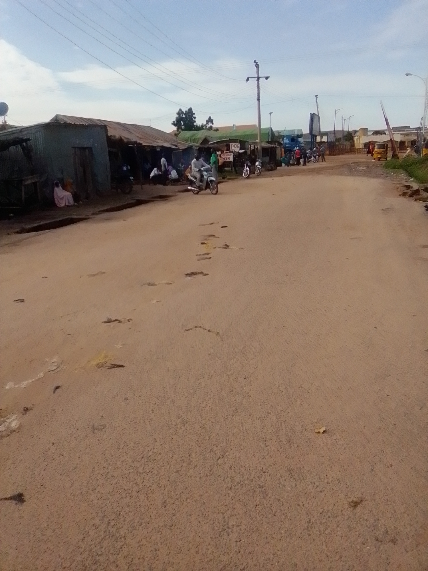 The image depicts a street scene in Gombe, Nigeria. On the left side of the road, there are several corrugated iron structures with tin roofs, resembling small shops or stalls. A woman in a pink hijab is seated outside one of these structures. A few other people are gathered around these stalls, and a motorcycle is parked nearby.
The road itself appears to be unpaved, with a sandy, reddish-brown surface. There are some potholes and uneven patches on the road. Further down the street, more buildings and a few more vehicles are visible, including a tricycle. A power pole with wires stretches across the sky. The overall impression is of a busy, informal commercial area within the city. The sky is cloudy, indicating a typical day in Gombe.