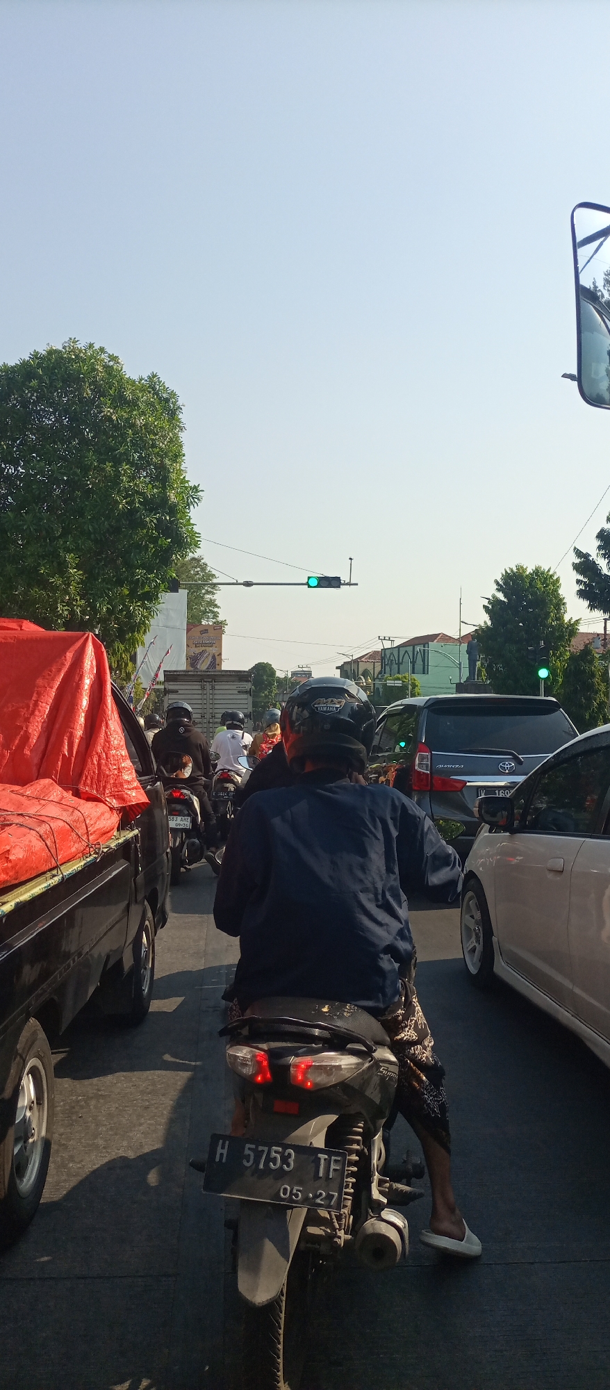 The image shows a traffic jam on a sunny day in Jati Kulon, Indonesia. The viewpoint is from behind a motorcycle rider, showing a line of vehicles including motorcycles, a truck with a red tarp, and cars. A traffic light in the distance is green. The road surface is asphalt. The rider is wearing a dark blue long-sleeved shirt and patterned pants. Another motorcycle is visible ahead with license plate H 5753 TF. Buildings and trees are visible in the background.