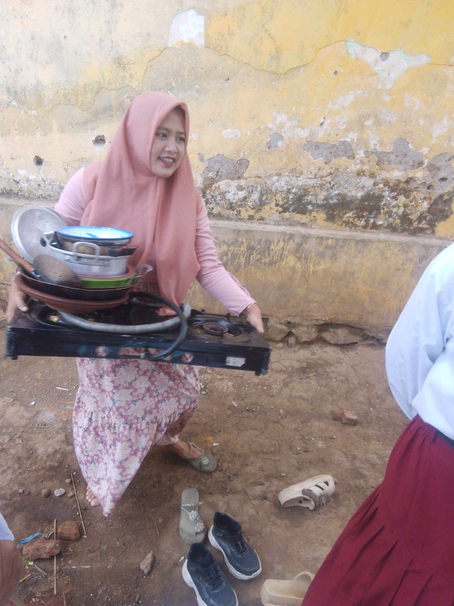 In Betek, Indonesia, a woman wearing a pink hijab and a floral patterned dress carries a stack of dishes and a portable stove. She is walking on a dirt ground in front of a weathered, yellow-painted wall. Nearby, two pairs of shoes are visible: a pair of black sneakers and a pair of light-colored sandals. A portion of another person, wearing a white shirt and a red skirt, is also visible in the foreground on the right side of the image. The environment appears to be an outdoor, somewhat dilapidated area.