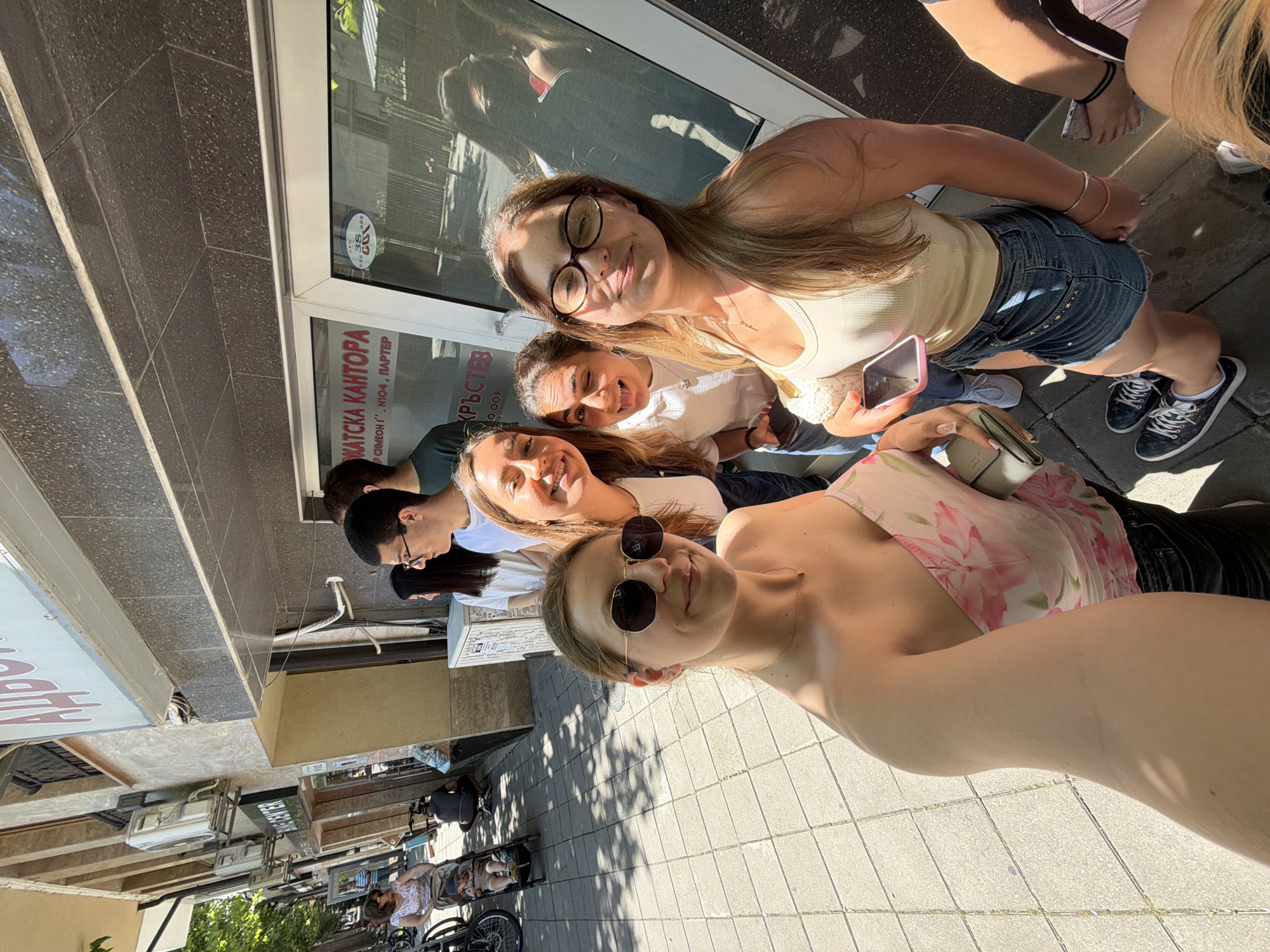 In Burgas, Bulgaria, four young women are standing on a sidewalk in front of a building entrance. The woman in the foreground on the left is taking a selfie, wearing sunglasses and a floral off-the-shoulder top. To her right, another woman is winking and sticking out her tongue, wearing a white t-shirt. A third woman, to the far right, is wearing glasses and a tank top, with her eyes partially closed. Behind them, other individuals are visible, including a man in a green t-shirt. The building facade features a sign in Bulgarian that appears to be for an office. The sidewalk is paved with square tiles, and trees cast shadows on the ground. Sunlight illuminates the scene, suggesting it is daytime. The overall atmosphere is casual and appears to be a group of friends posing for a photo.