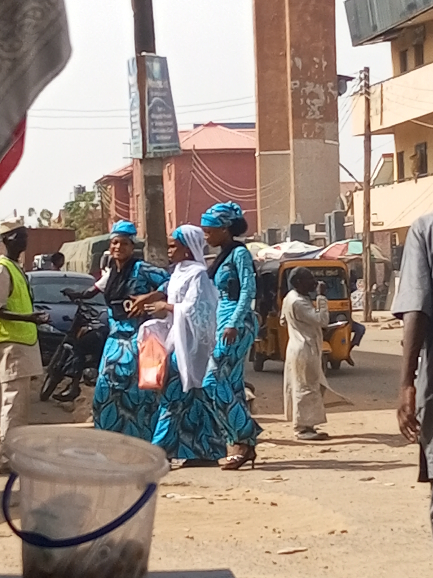 A busy street scene in Maiburiji, Nigeria. Two women in blue patterned dresses and matching headwraps walk alongside a woman in a white headscarf. All three are on a sandy ground road. To the left, a man in a yellow vest appears to be talking to the women. A motorcycle and a car are nearby. In the background, there's a yellow tricycle taxi, buildings, and utility poles. Other individuals are scattered around the scene. In the foreground, a plastic bucket and a person in gray are partially visible.