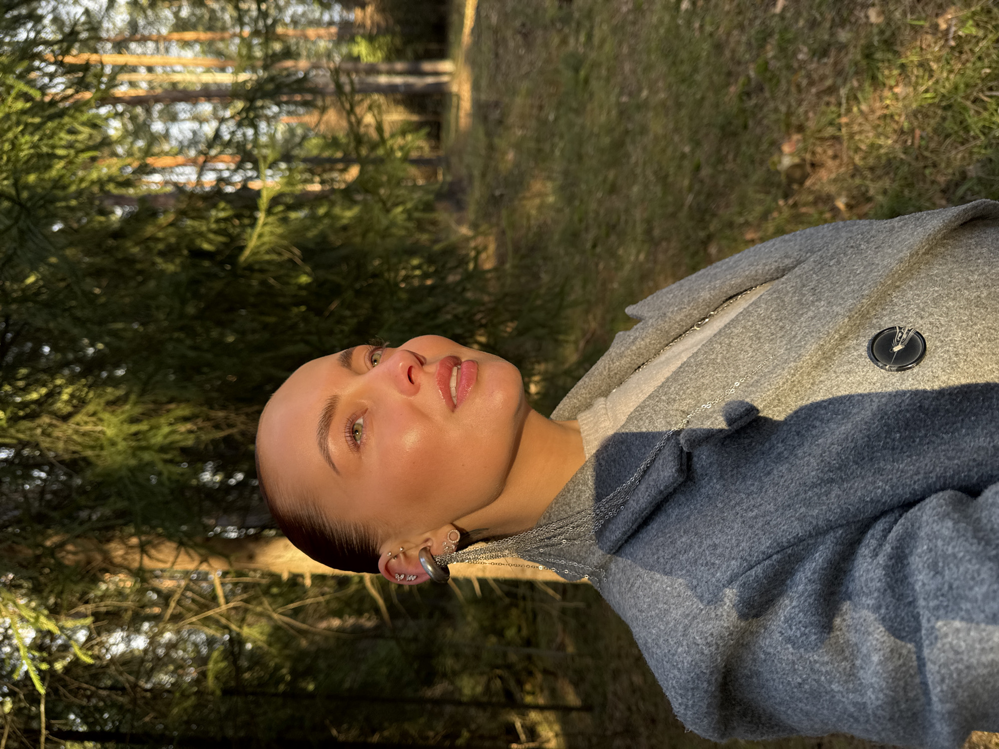 The image shows a woman outdoors in a wooded area, likely in Прикамский, Russia, bathed in warm, golden light suggesting late afternoon or early morning. The woman, from her torso upwards, is looking upwards and slightly to her left with a serene expression. Her light skin is illuminated, highlighting her green eyes and red lipstick. Her hair is pulled back, revealing multiple silver piercings in her left ear, including a large hoop and several smaller studs. She is wearing a grey wool coat, with a silver chain necklace visible beneath. A dark, circular badge or pin with a lighter, abstract symbol (possibly a stylized "V" or wings) is affixed to her coat's lapel. The background features dense evergreen trees, with sunlit patches of grass and foliage on the forest floor. The scene is tranquil, with dappled light and shadows created by the sunlight filtering through the trees. No legible text is visible in the image.