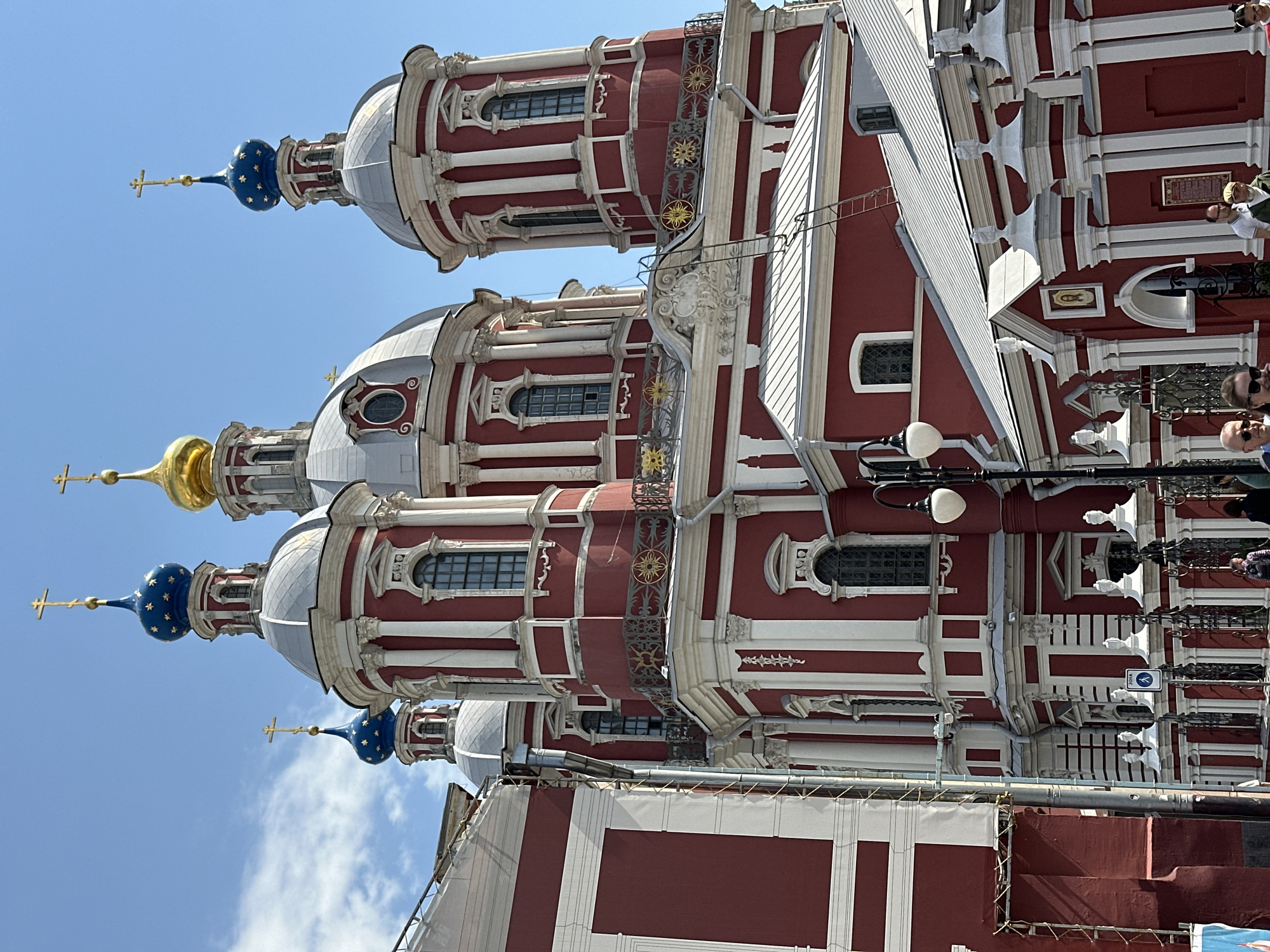 The image shows the exterior of the Church of St. Nicholas in Khamovniki, Moscow, Russia. The church features red and white striped walls and multiple domes, some topped with blue, gold, and silver colors. The domes are adorned with crosses. The sky is blue, and some people are visible in the background. Scaffolding and other temporary structures appear around the base of the building, indicating ongoing maintenance or construction.