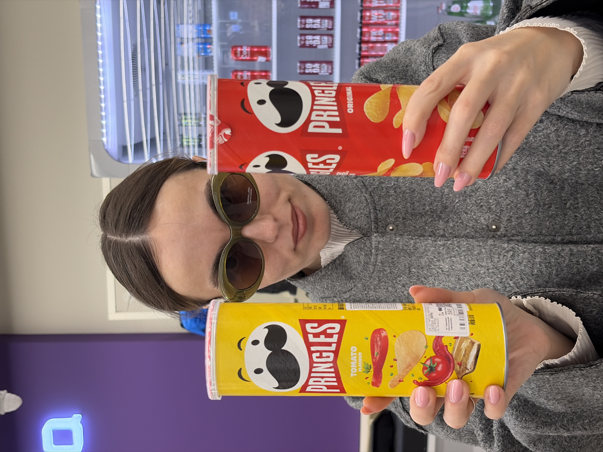 A woman wearing sunglasses and a grey coat is holding a can of yellow Pringles Tomato Flavor chips and a can of red Pringles Original chips in an indoor location. A refrigeration unit with other snack chips is behind her. A purple wall with an illuminated blue block is in the lower left of the frame. The price tag on the can of Pringles Tomato Flavor chips shows the Russian city of Izhevsk.
