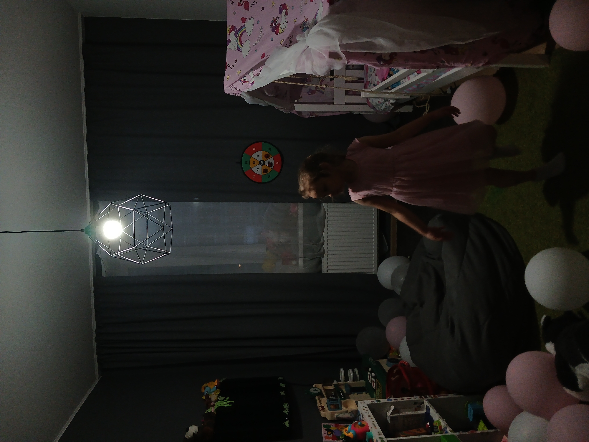 The image shows a dimly lit bedroom, possibly in Yekaterinburg, Russia, where a young girl in a pink dress is standing. The room is decorated with balloons of various colors and a unicorn-themed bed canopy. A large, dark beanbag chair is in the center of the room, and shelves filled with toys are visible on the left side. A dartboard hangs on the wall, and a modern geometric pendant light fixture illuminates the space from above.