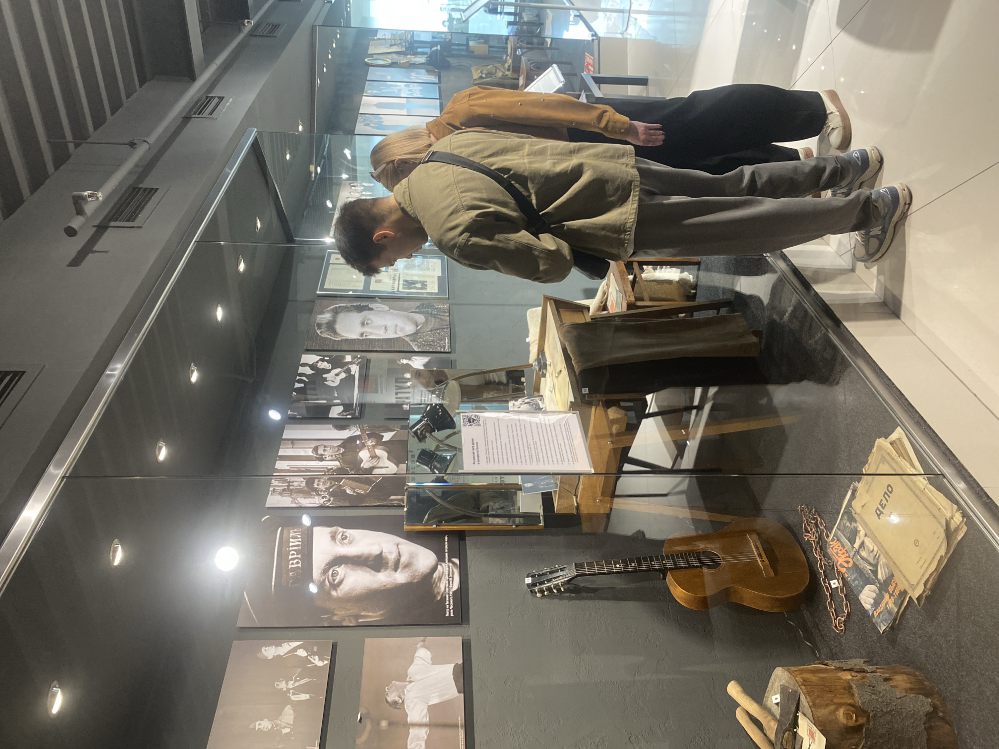 The image shows the interior of a museum exhibition, likely dedicated to the life and work of Vysotsky, given the prominent display of his image and a guitar. Two individuals, a man and a woman, are observing the exhibition. The man is wearing a green jacket and grey pants, while the woman is in a brown top and black pants. They are positioned behind a glass display case containing a guitar, a wooden stump with tools, and various documents and magazines. The exhibition features black and white photographs on the wall, including portraits and group shots. The environment appears to be a modern museum with track lighting and clean lines. The text on the signs is in Russian. The provided information indicates the museum is located in Yekaterinburg, Russia.