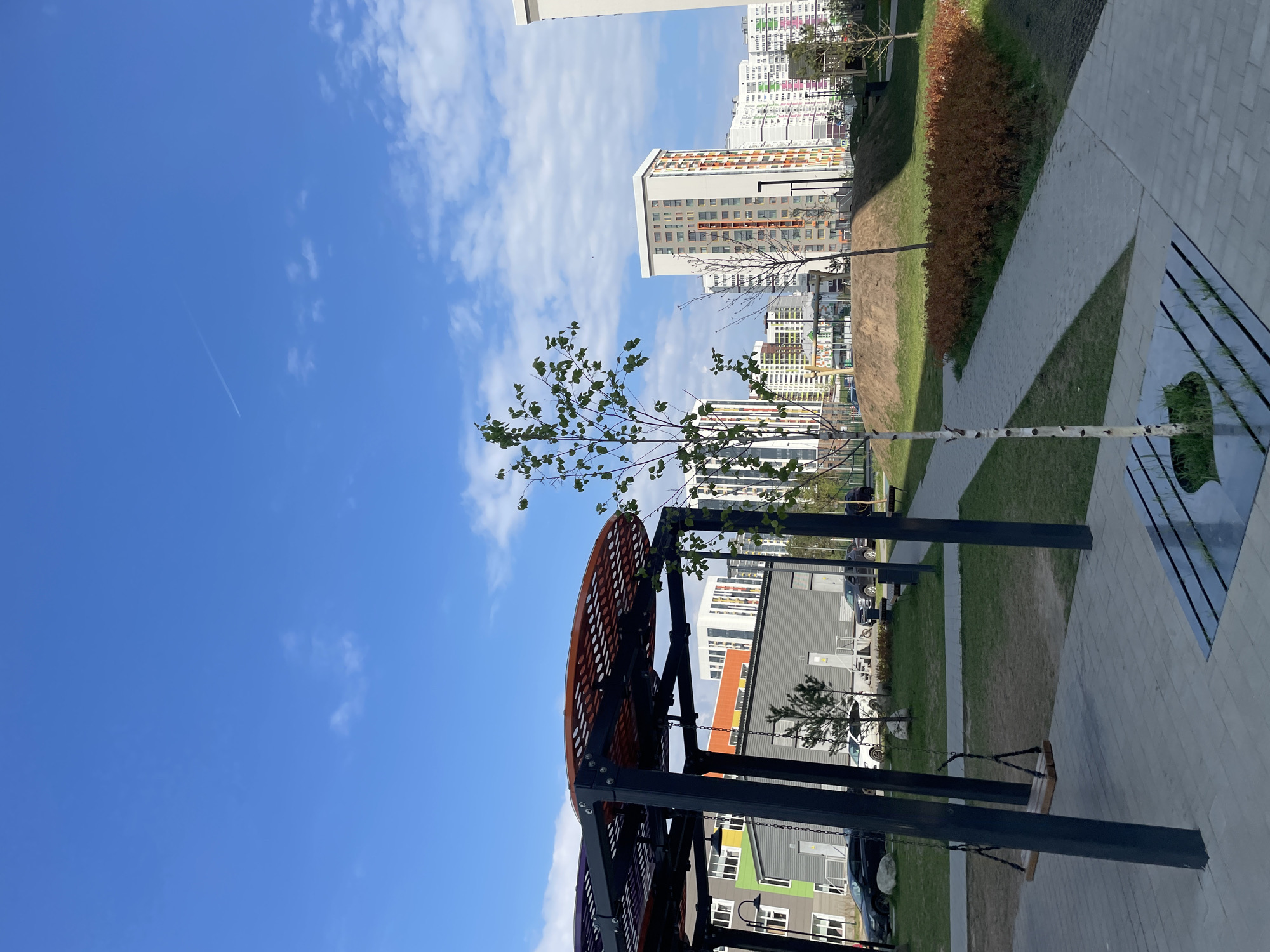 The image shows an urban landscape, likely Izhevsk, Russia, with a focus on a modern park area. In the foreground, a playground structure with a large, circular orange-colored component and swings is visible. A young birch tree stands near the playground. Beyond the park, multiple tall residential buildings, painted in various pastel colors, form the cityscape backdrop. The sky is blue with scattered clouds and a contrail from an aircraft. The park area includes grassy spaces and paved walkways.