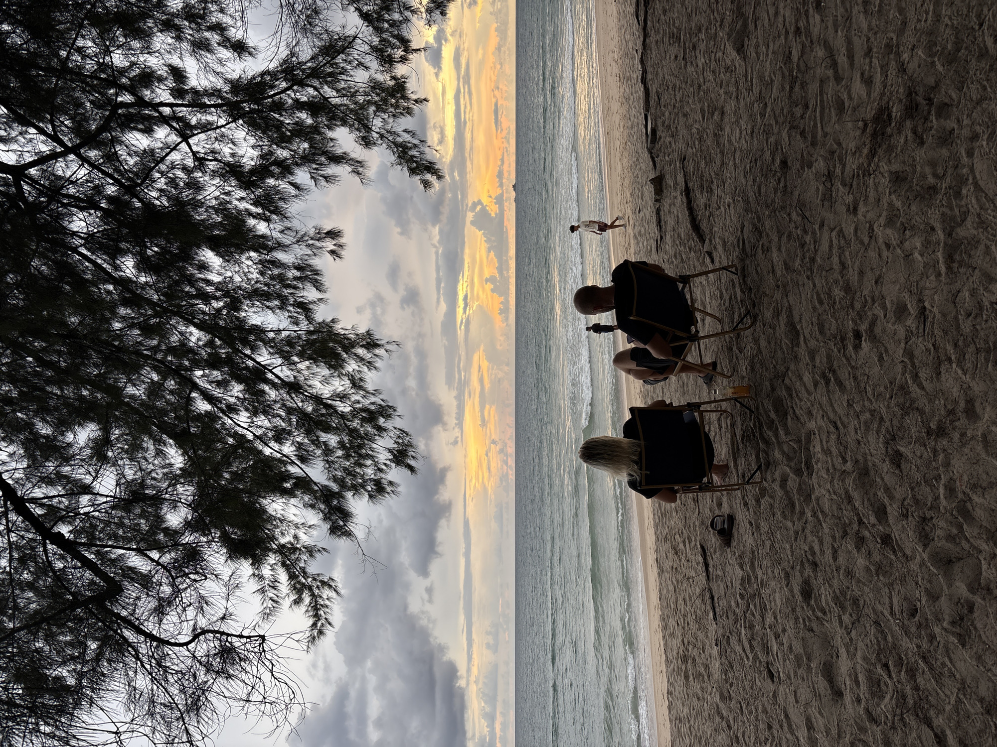 A view of the beach in Choeng Thale, Thailand, at dusk. Two people are seated in chairs on the sand, facing the ocean. The sky is cloudy with hints of orange and yellow from the setting sun. Waves gently lap the shore, and a few other figures are visible walking on the beach in the distance. The foreground is dominated by the textured sand and the silhouette of pine tree branches overhead.