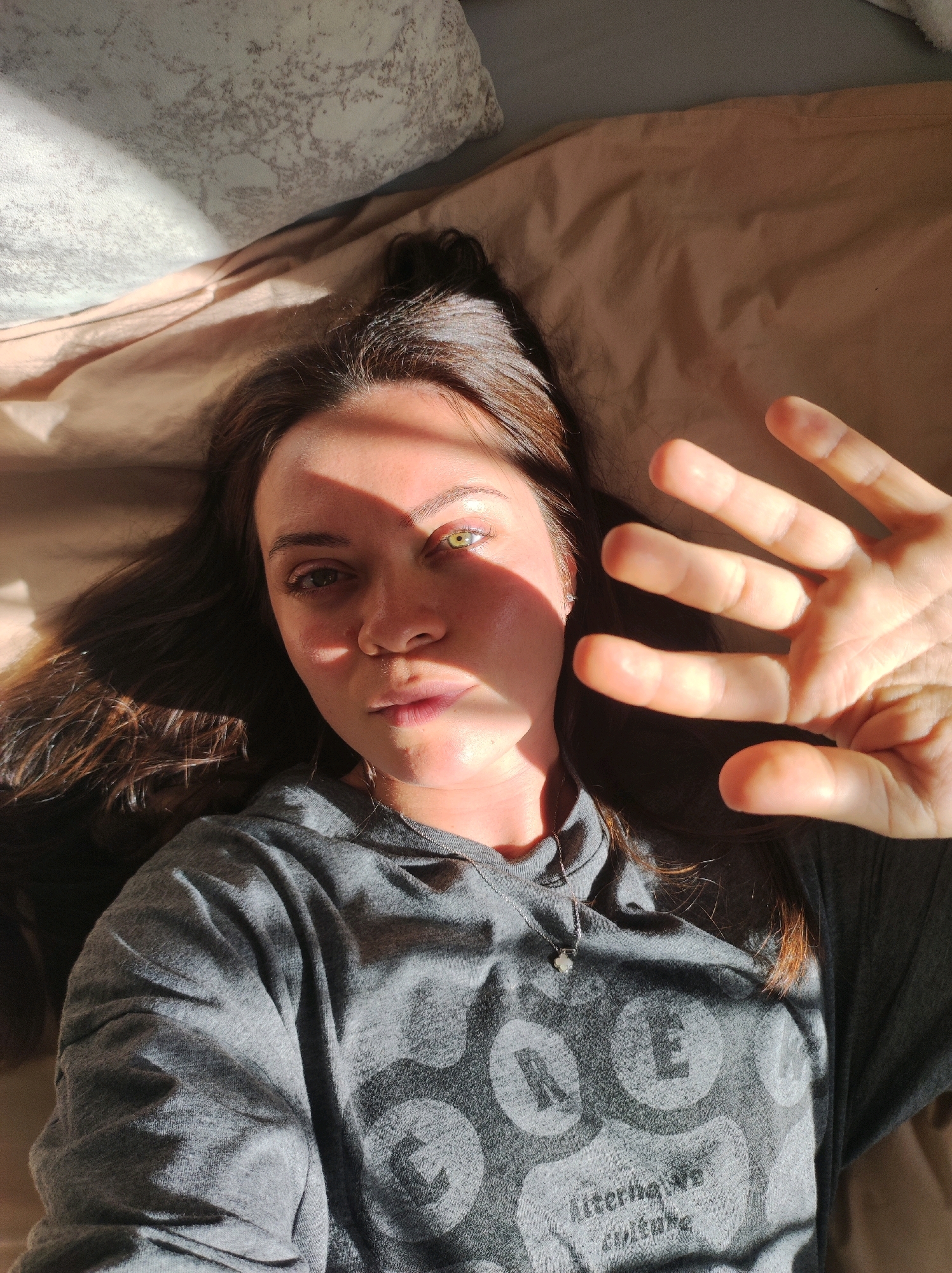 A young woman with long, dark hair lies on her back on a bed with wrinkled, light brown sheets. Sunlight streams in from the left, casting a shadow across the left side of her face and illuminating the right side. Her right eye appears to be a different color, possibly due to the light. She is wearing a grey hooded sweatshirt with a circular pattern of letters and the words "alternative culture" printed on it. Her right hand is raised, obscuring part of her face. A small, silver necklace with a clover-shaped pendant is visible on her chest. The background consists of a pillow on the left and more bedding. The image appears to be a selfie taken indoors. No specific location in Chaikovsky, Russia, can be identified from the visual information provided.