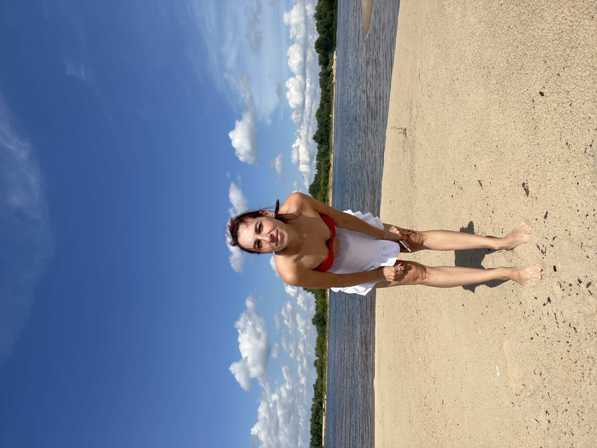 A full-shot image taken from a low angle captures a young woman on a sandy beach under a bright blue sky with scattered white clouds. She is wearing a red bikini top and a white skirt. The woman is leaning forward, with her hands resting on her knees, and looking towards the camera. Her feet are bare and planted on the sand. Behind her, a wide river stretches across the frame, with a lush green forest lining the opposite bank. The scene suggests a warm, sunny day at a waterfront location in Nizhny Novgorod, Russia.