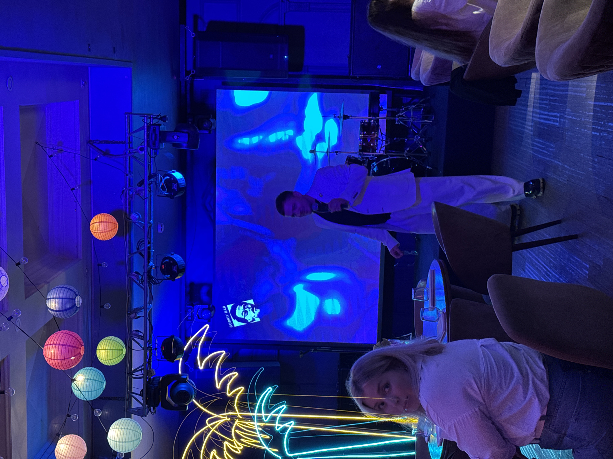 The image shows an interior space, possibly a bar or club, likely in Izhevsk, Russia. A man stands on stage, wearing a white suit and holding a microphone, seemingly performing. Behind him is a large screen displaying a blue abstract image with a smaller image of a man's face and the words "GORKY BAR". The stage includes a drum set and lighting equipment. In the foreground, a blonde woman looks at the camera. A brightly lit neon sign depicting a palm tree is also in the foreground. Paper lanterns hang from a lighting rig on the left side of the frame. The room is dimly lit with a blue hue. Tables and chairs are visible on the right side of the frame. A seated person is visible in the top right corner.