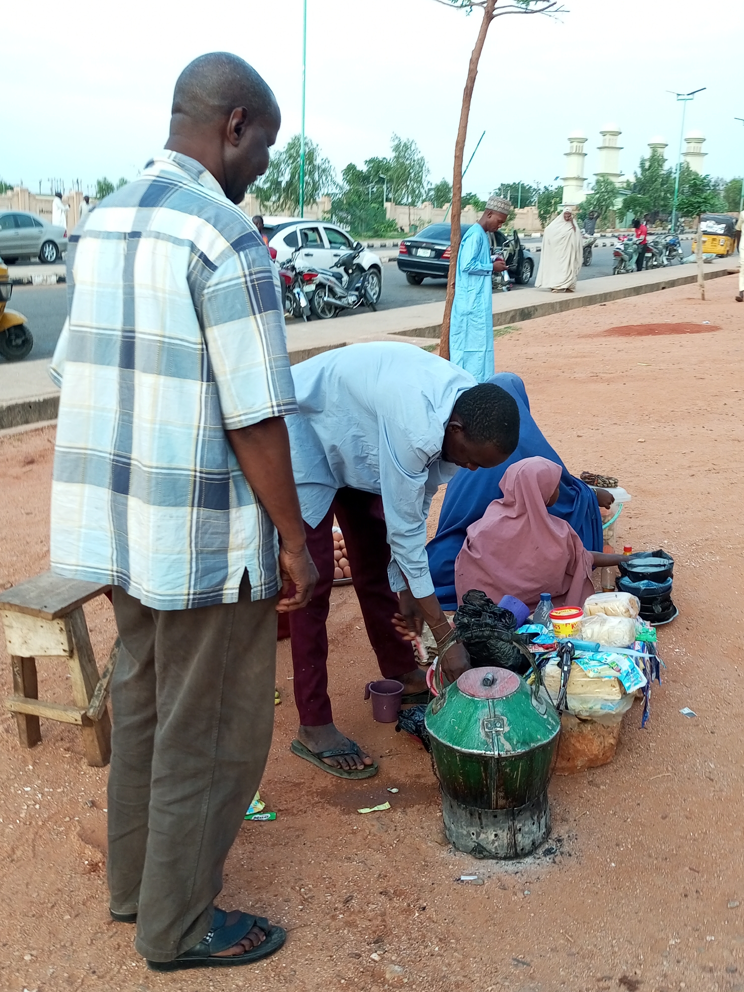 In the city of Gombe, Nigeria, a scene unfolds on a dusty, reddish-brown street with a few vehicles and people present. In the foreground, a man in a plaid shirt and brown pants stands facing away from the camera. He is looking down at a market stall where a woman in a pink hijab and a blue garment is seated. The stall displays various items, including food items, cooking pots, and what appears to be a large green kettle on a stove. Another man in a light blue shirt is bent over the stall, interacting with the woman. In the background, several cars, motorcycles, and a tricycle are visible on the road. Buildings with white domes, possibly a mosque, are seen in the distance. The overall impression is of a busy street market scene.
